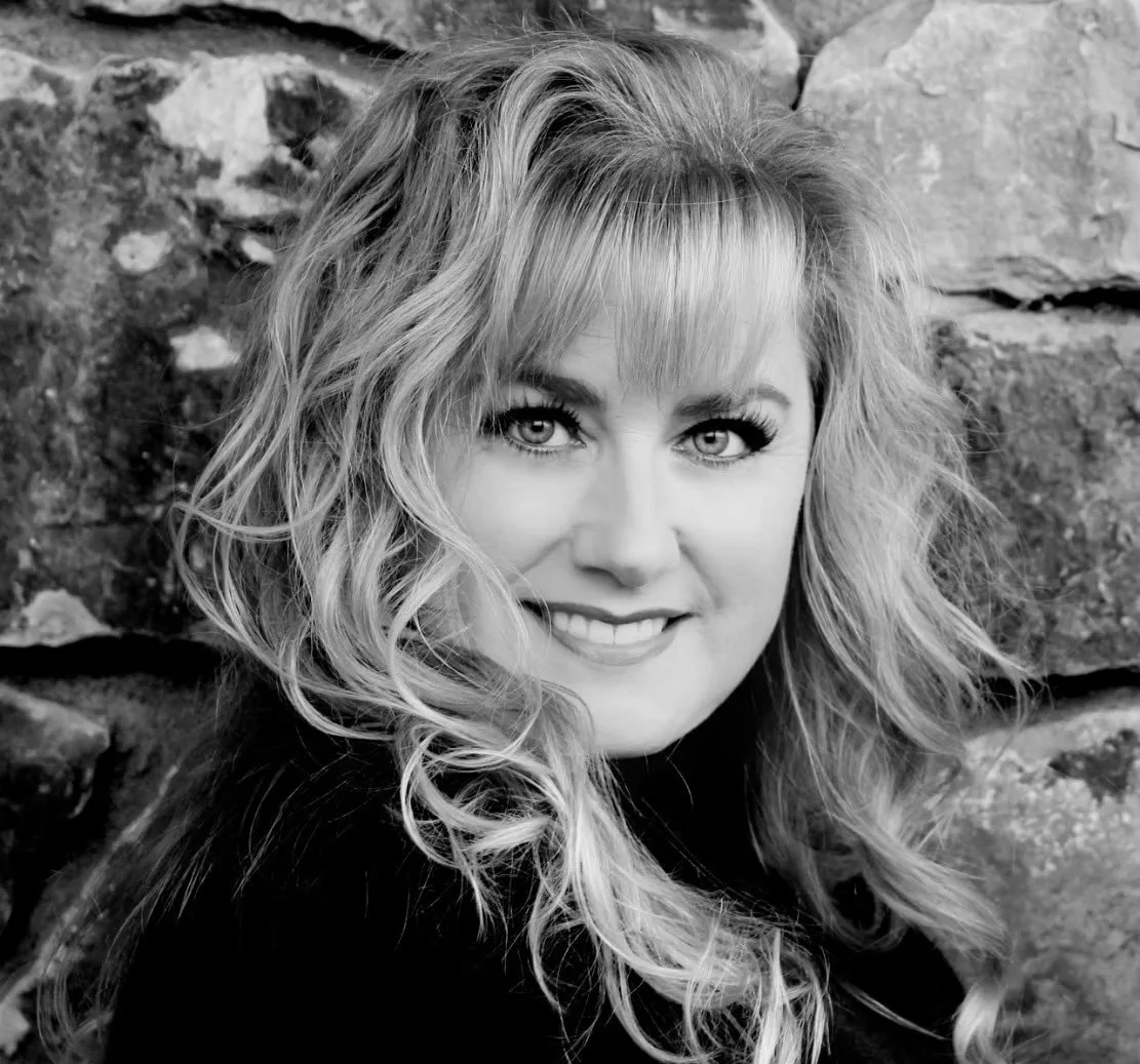 Black and white close-up photo of a smiling woman with wavy hair, wearing a dark top, standing against a stone wall. Downtown Novato Board Member.