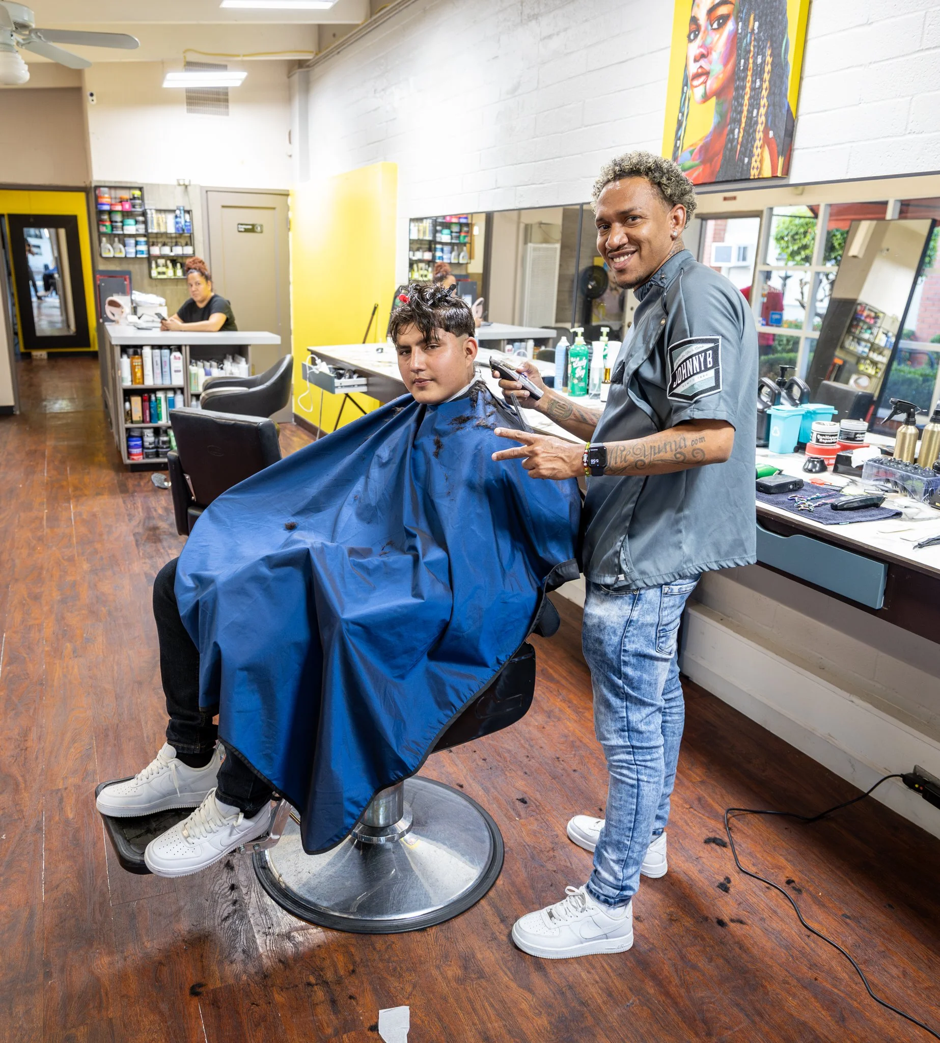 A barber giving a haircut to a young man in a salon in Downtown Novato California. The barber is smiling and holding a pair of scissors, while the young man is seated with a blue cape around him. 