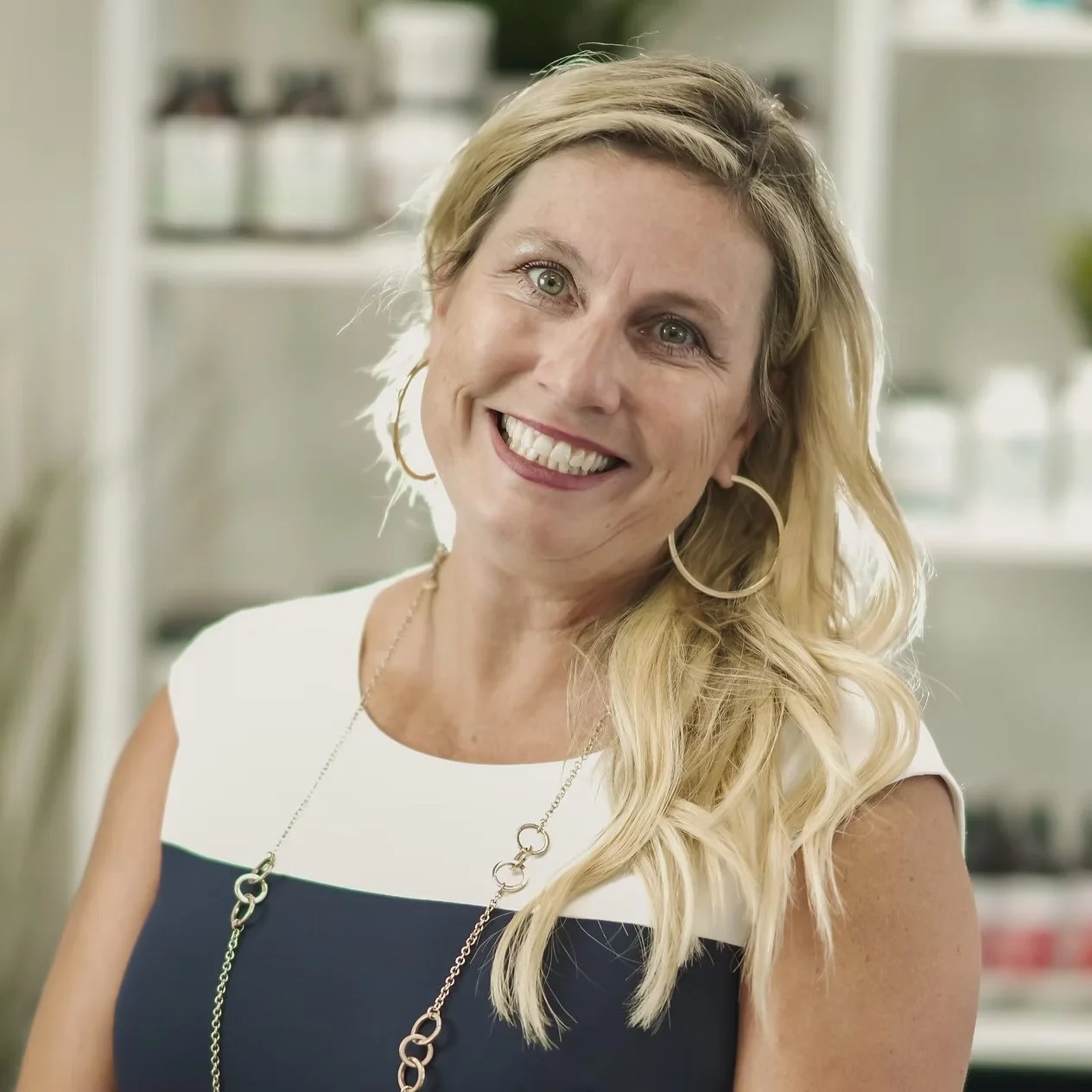 A smiling woman with long blonde hair, wearing large hoop earrings and a white and navy top, standing in a brightly lit indoor setting. Downtown Novato Board Member.