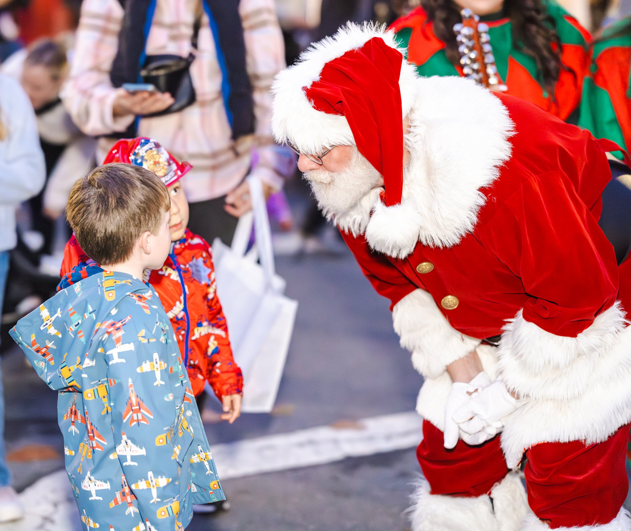 A person dressed as Santa Claus bending down and engaging with two children during a holiday event, with several onlookers in the background, in Downtown Novato California.