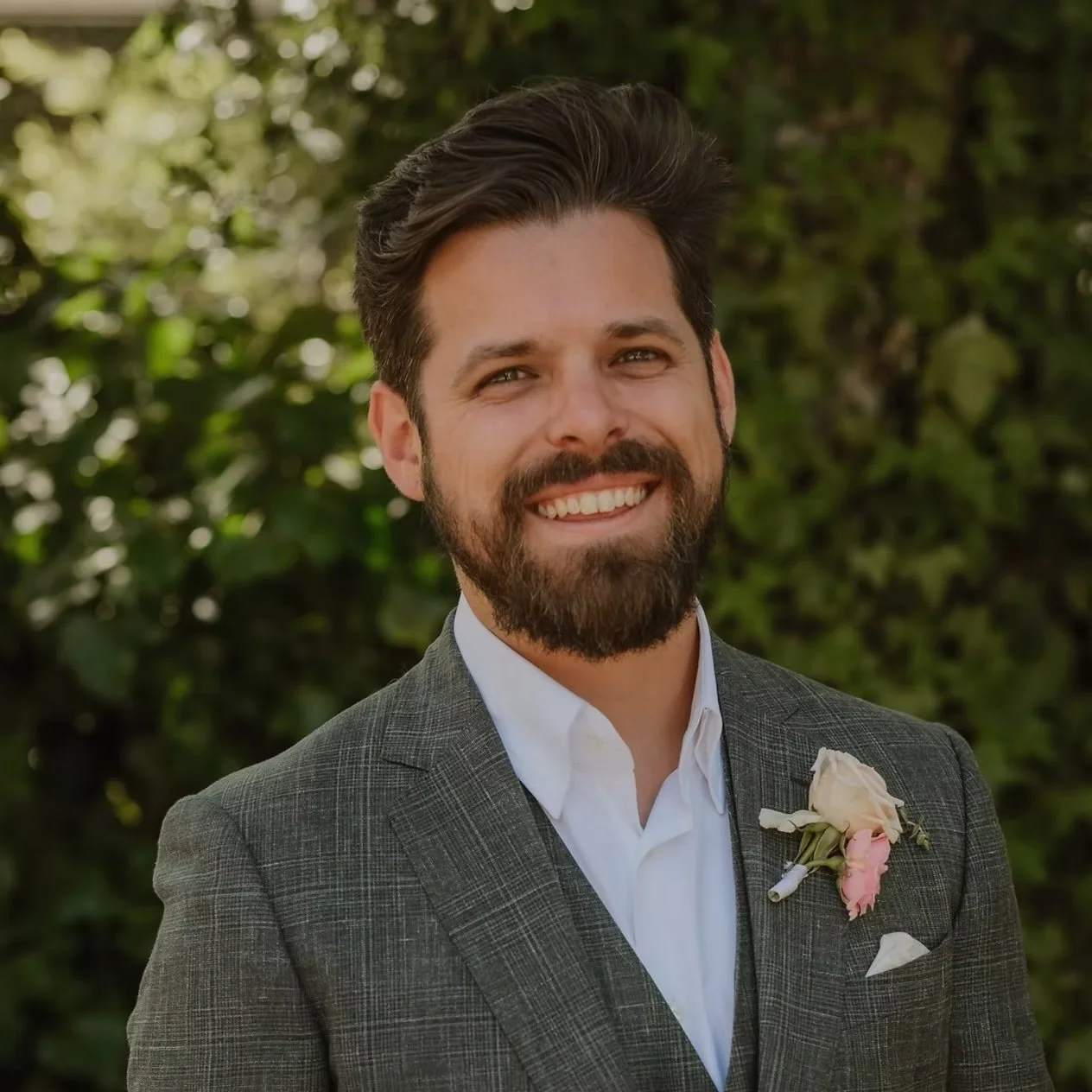 A smiling man with dark hair and a beard, wearing a gray plaid suit with a white shirt and a boutonniere, standing outdoors in front of green foliage. Downtown Novato Board Member.