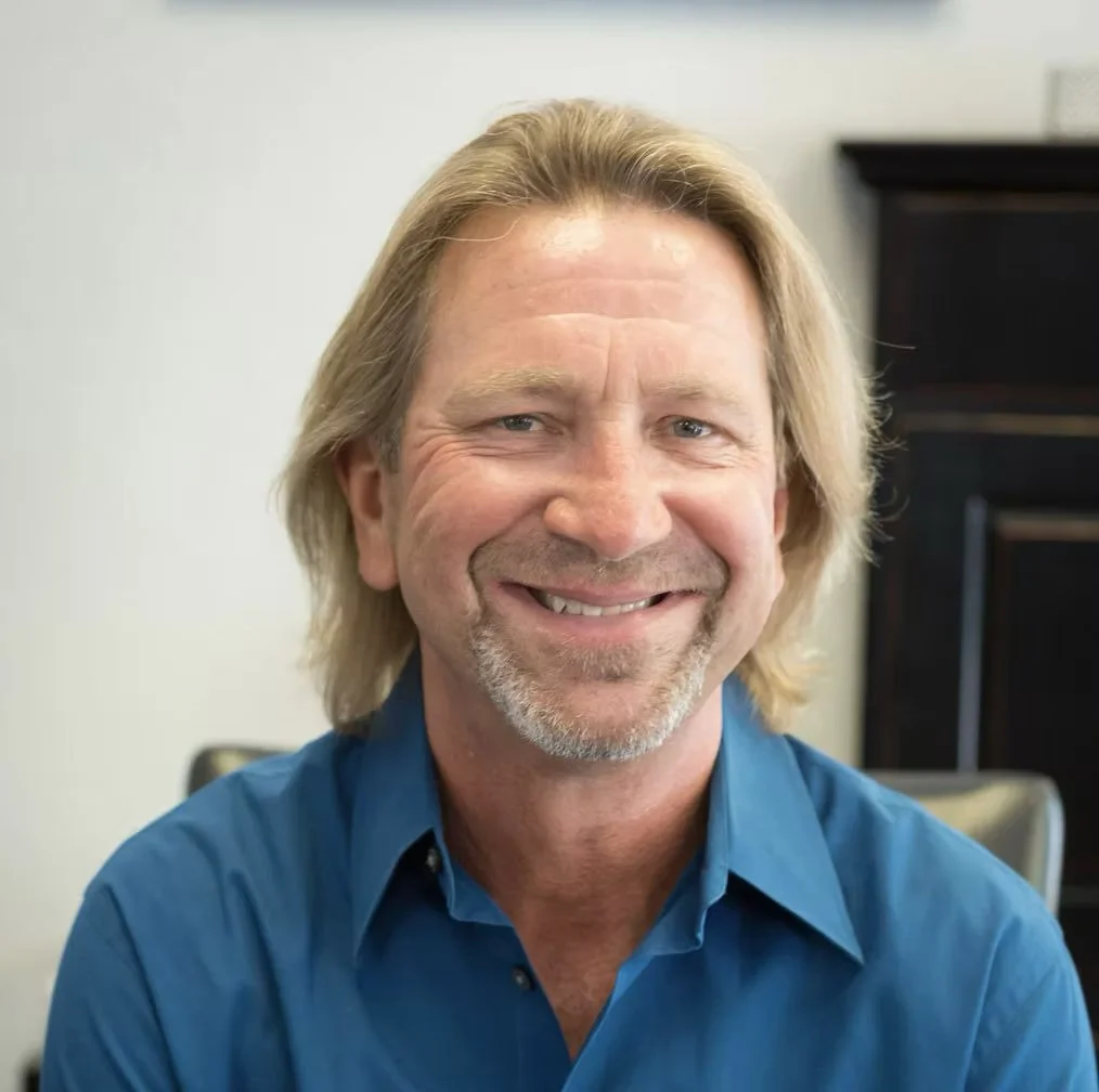 Portrait of a middle-aged man with shoulder-length blond hair, smiling, wearing a blue shirt, in an office environment. Downtown Novato Board Member.
