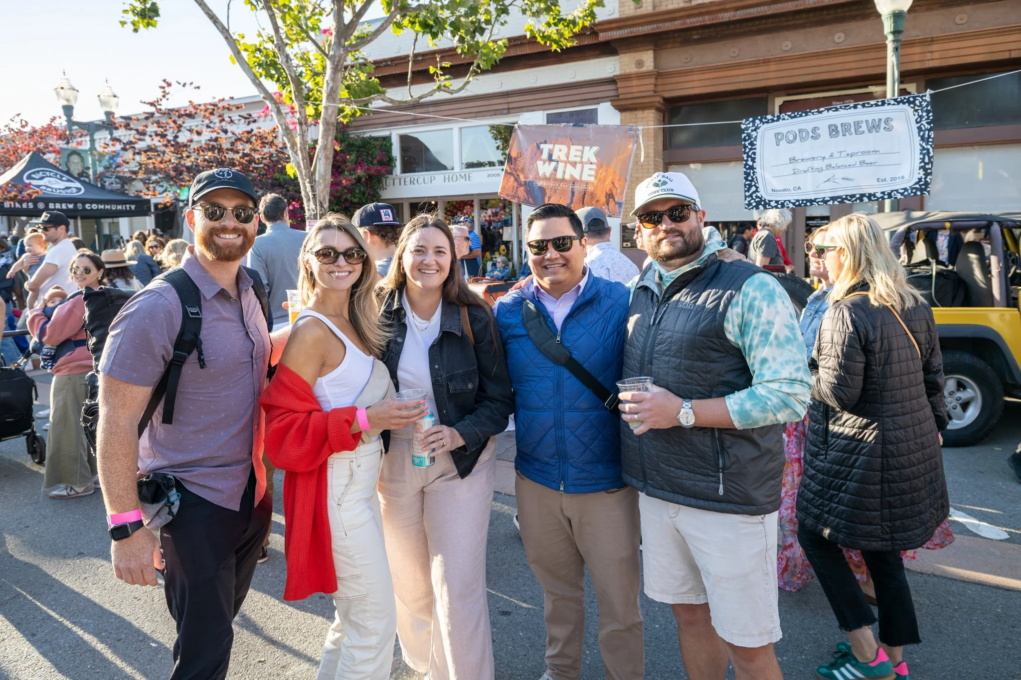 A group of six young adults smiling and posing together outdoors during a festival or fair, with booths and crowd in the background, in Downtown Novato California.