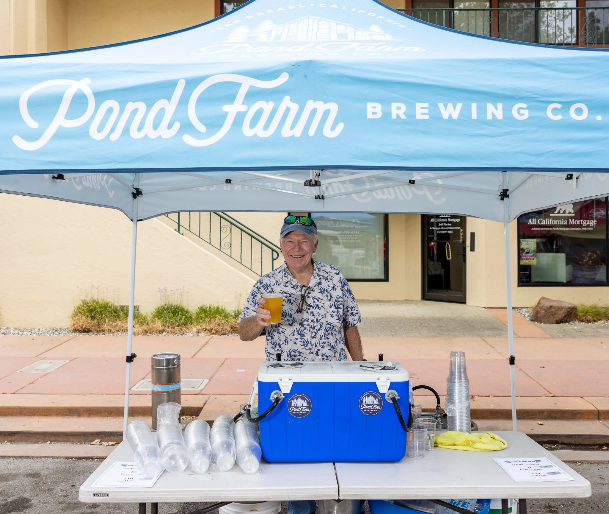 A smiling older man in a blue and white floral shirt holding a yellow drink stands behind a table at an outdoor event, in Downtown Novato California.