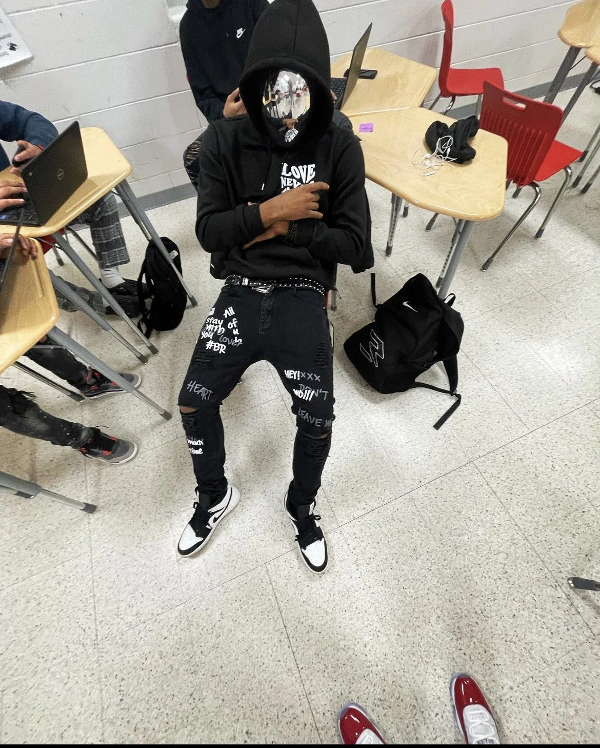 A person wearing a black hoodie with the hood up, a black face mask, and black jeans with white graffiti-style writing, standing in a classroom near desks, with their arms crossed and pointing at themselves with their right hand.