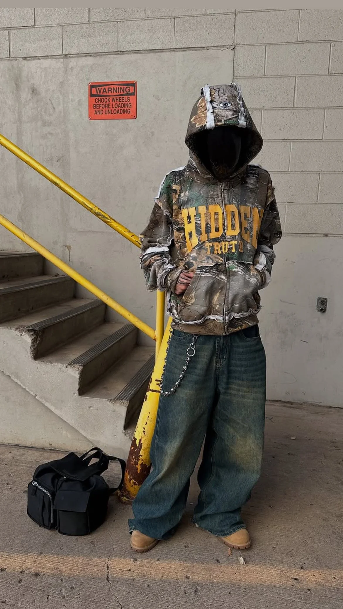 A person wearing a camouflage hoodie with the words "Hidden Trout" in yellow, baggy jeans, and tan work boots, standing next to a yellow handrail on a stairwell, with a black bag on the ground near them.