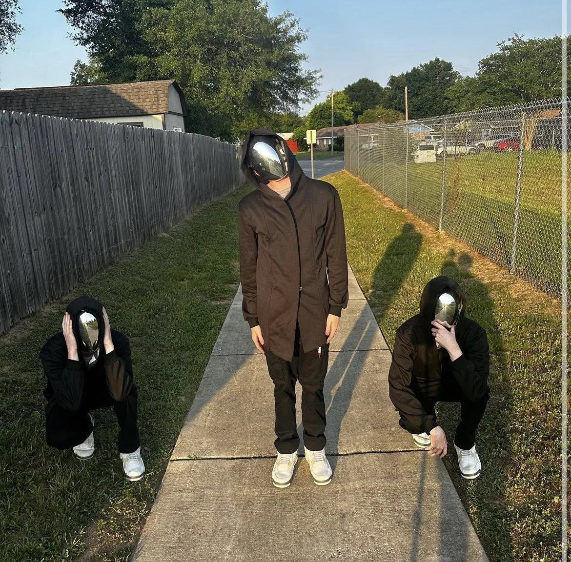 Three individuals wearing black jackets, black pants, white sneakers, and reflective helmets on a sidewalk with grass on both sides.