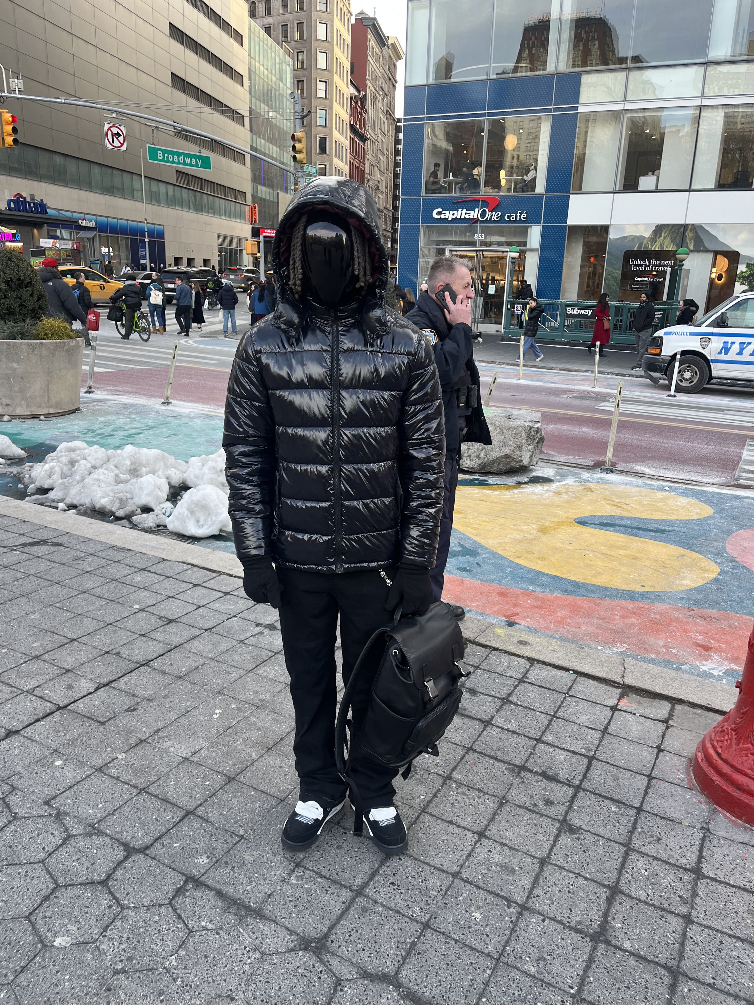 Person wearing a black shiny puffer jacket, black pants, black gloves, black shoes with white accents, and carrying a black backpack standing on city sidewalk with snow patches, next to a red fire hydrant, with City police and city buildings in the b