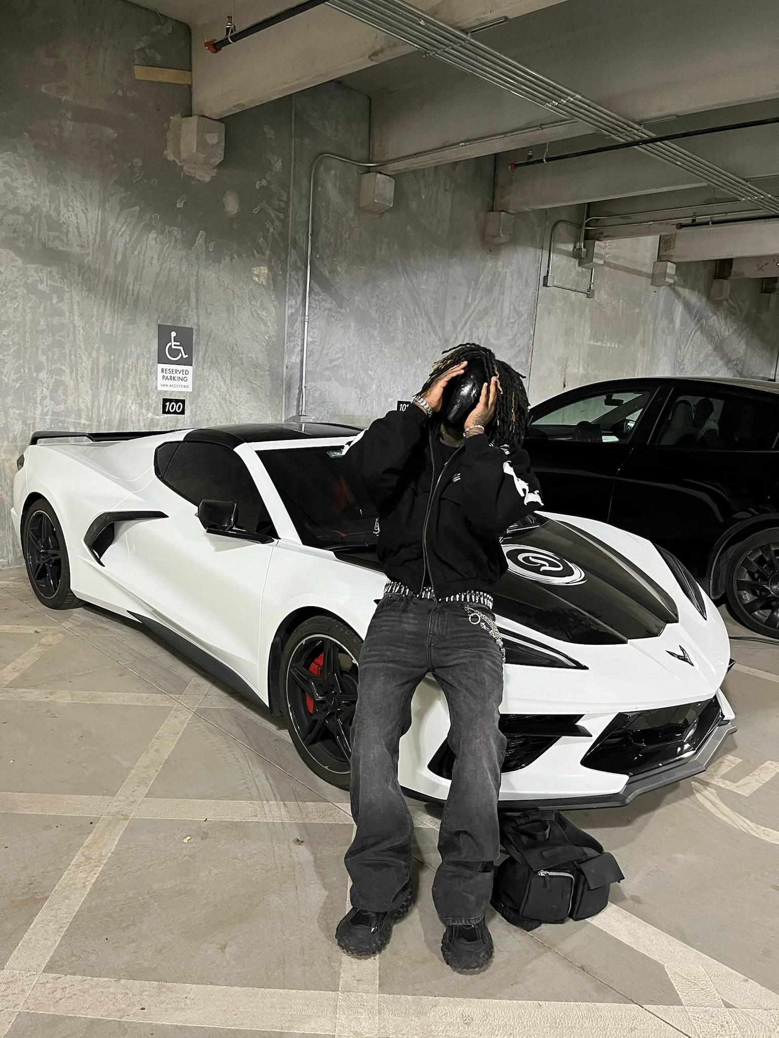 Person standing in front of a white sports car with black accents in a parking garage, holding a helmet over their head, with a black backpack on the ground nearby.
