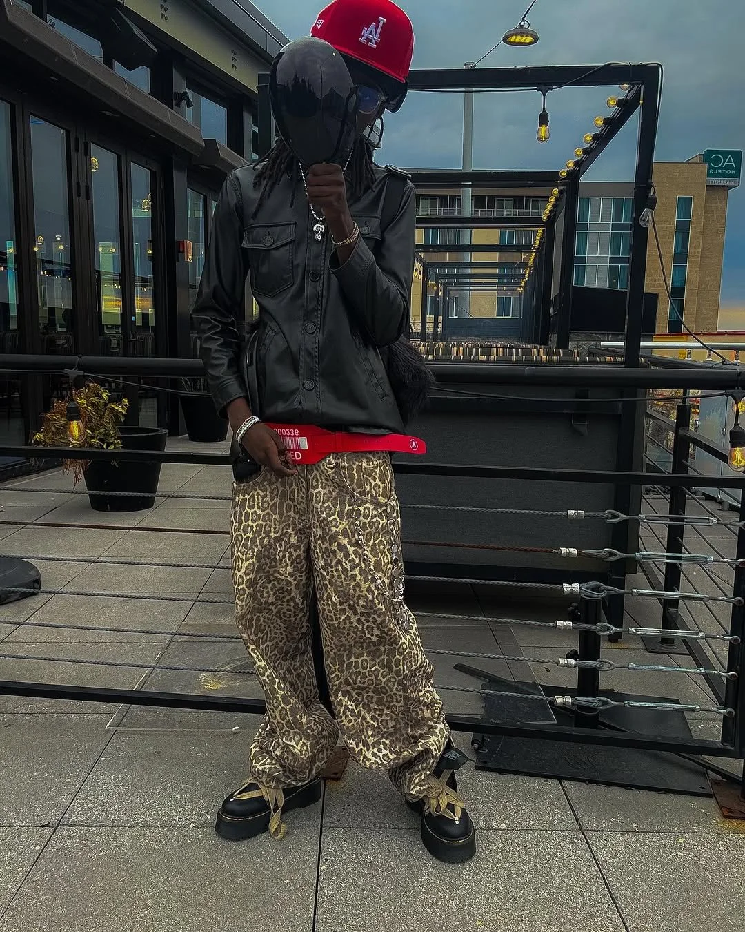 Person wearing a black leather jacket, leopard print pants, black shoes with yellow laces, red baseball cap, and holding a red ticket, standing outdoors on a balcony.