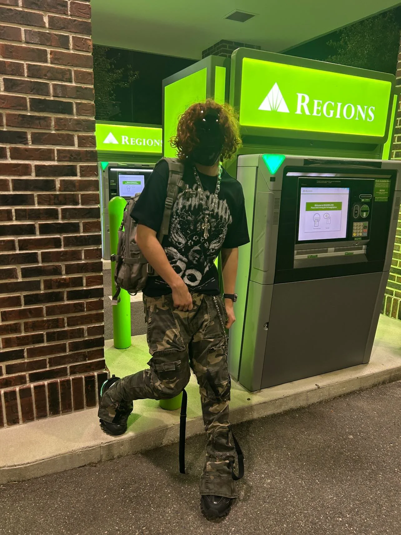 A person with a prosthetic leg standing next to an ATM at night outside a Regions bank branch, wearing a black mask, a graphic T-shirt, camouflage pants, and carrying a backpack and accessories.