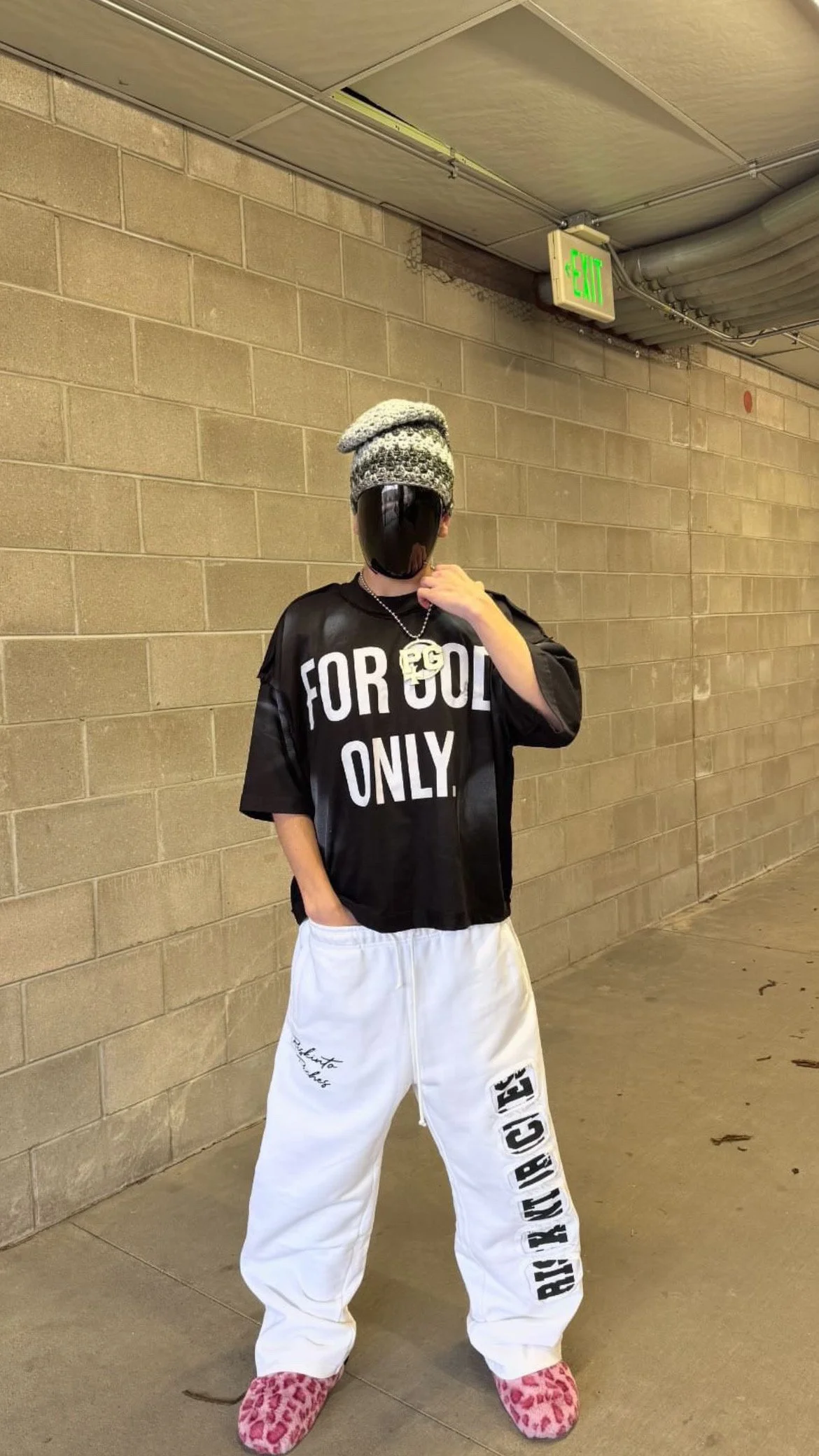Person standing in an indoor hallway wearing a faceless mask, a knit hat, a black T-shirt with white text "FOR GOD ONLY," white sweatpants with black text on one leg, pink leopard print slippers, and a chain necklace, with an green exit sign above.