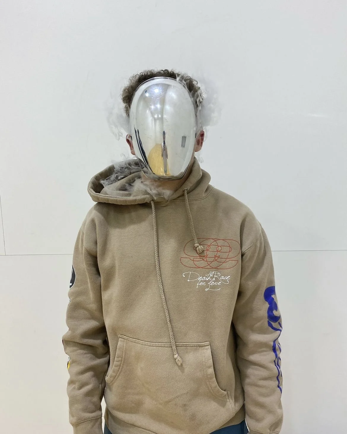 Person wearing a beige hoodie with text and patterns, with a reflective metallic mask covering their face, standing against a plain white wall.