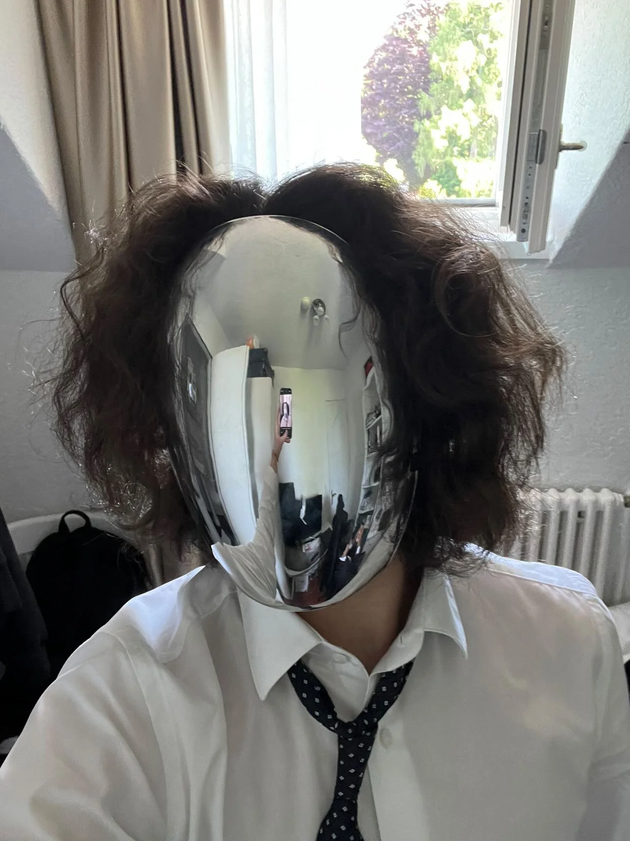 Person wearing a reflective mask, white shirt, and polka-dot tie, taking a selfie in a mirror with an open window and a radiator visible in the background.