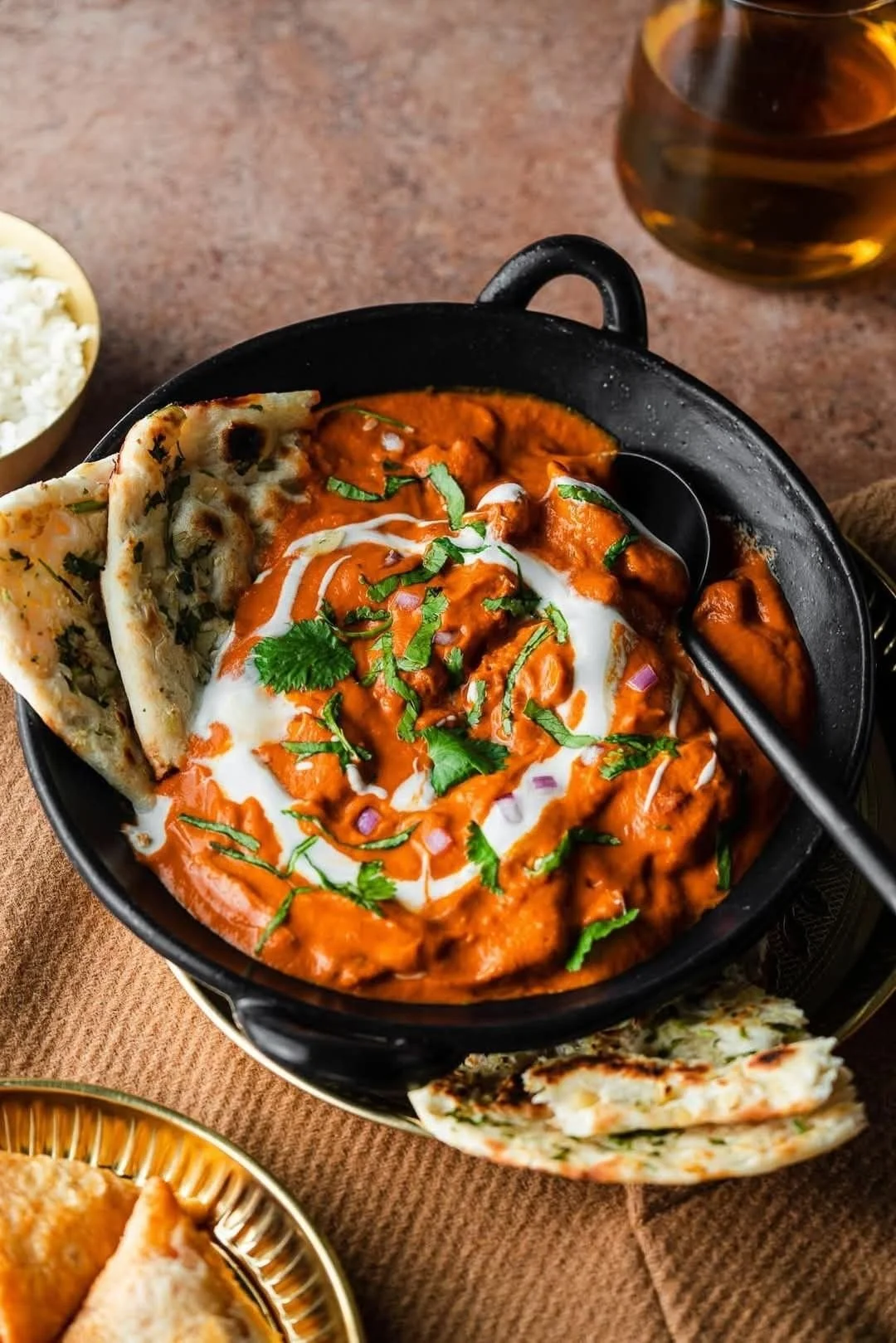Indian dish of butter chicken garnished with cilantro and served with naan bread. A bowl of rice and a glass of dark beverage are partially visible in the background.