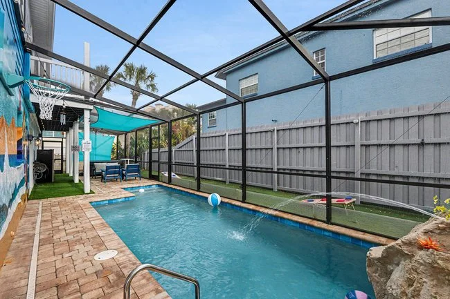 Enclosed backyard pool area with a small rectangular pool, brick pavement, and basketball hoop, surrounded by a screen enclosure and a gray wooden fence, with nearby houses and palm trees in the background.