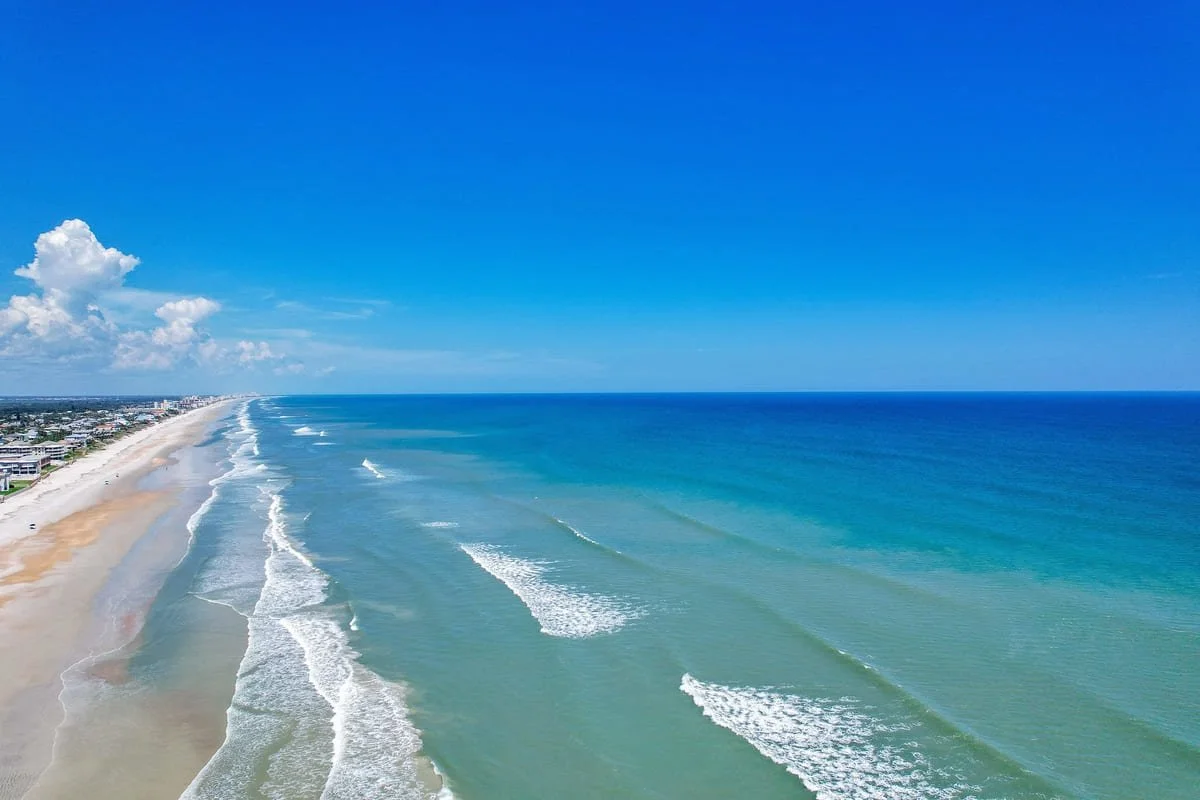Aerial view of a sandy beach along the ocean with waves, blue sky, and a few clouds.
