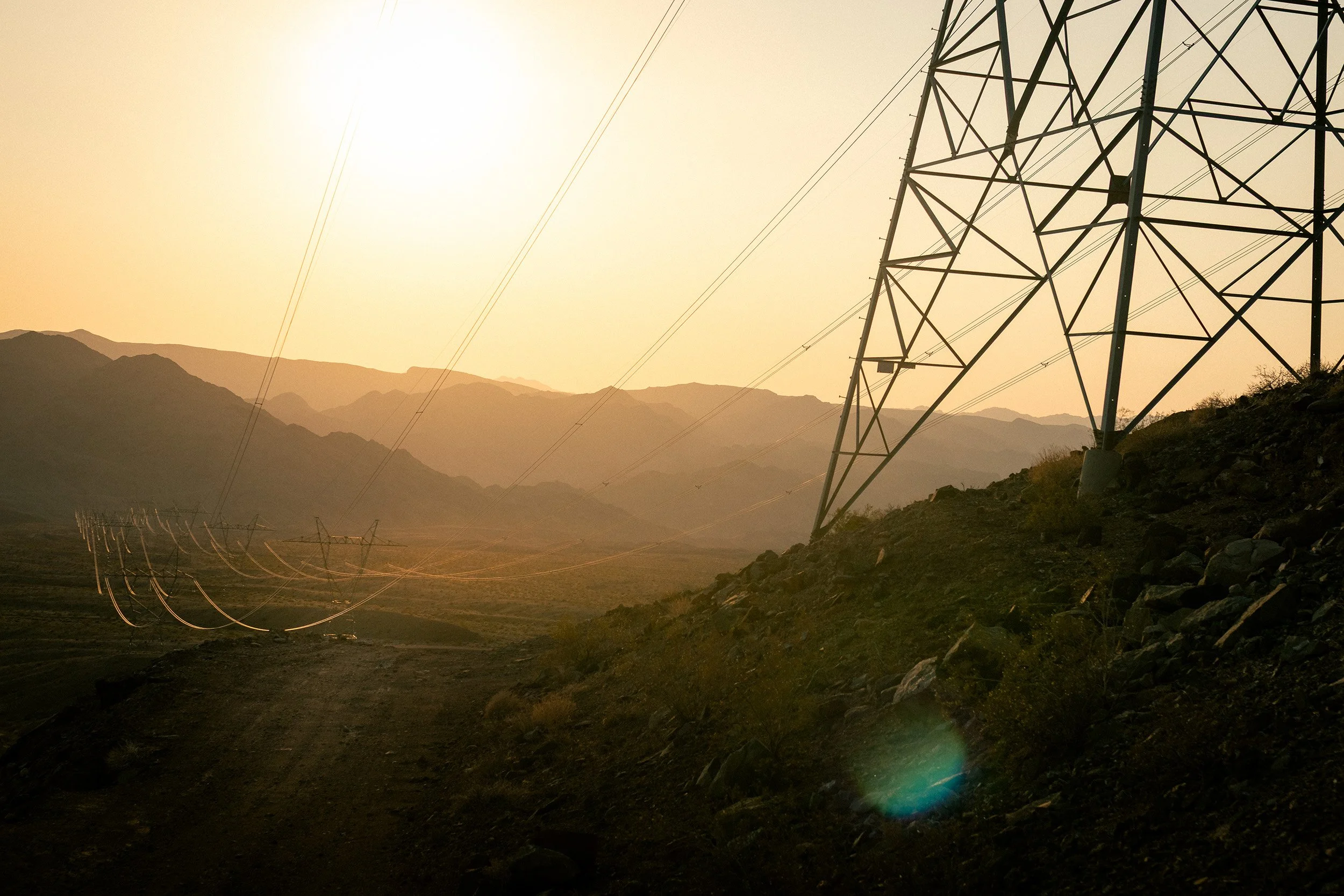 Power lines and transmission towers on a hillside at sunset with mountains in the background.