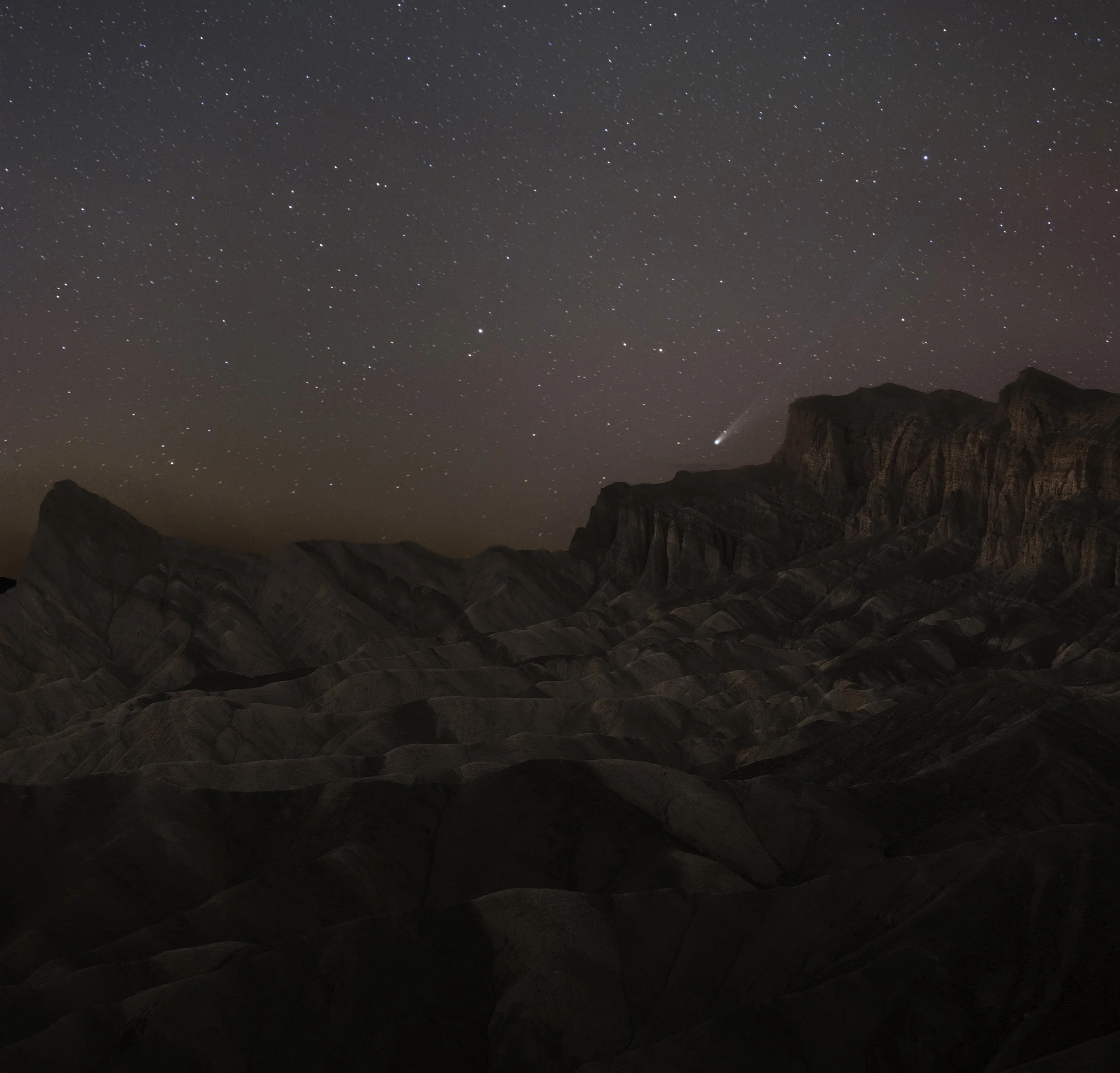 Nighttime landscape of rugged, layered mountains under a starry sky with a comet visible in the upper right.