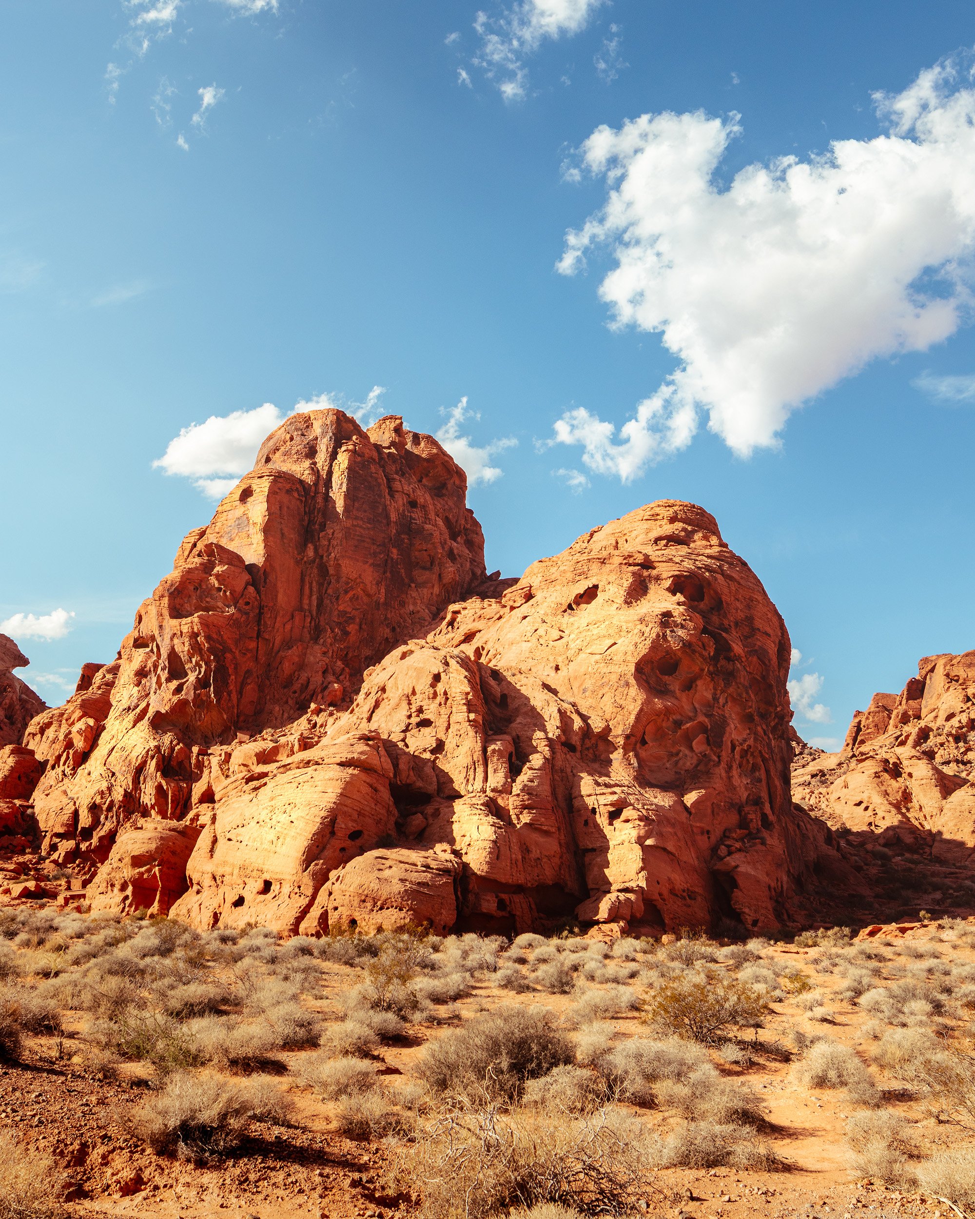 Large red and orange rock formations in a desert landscape with dry bushes, under a blue sky with scattered white clouds.