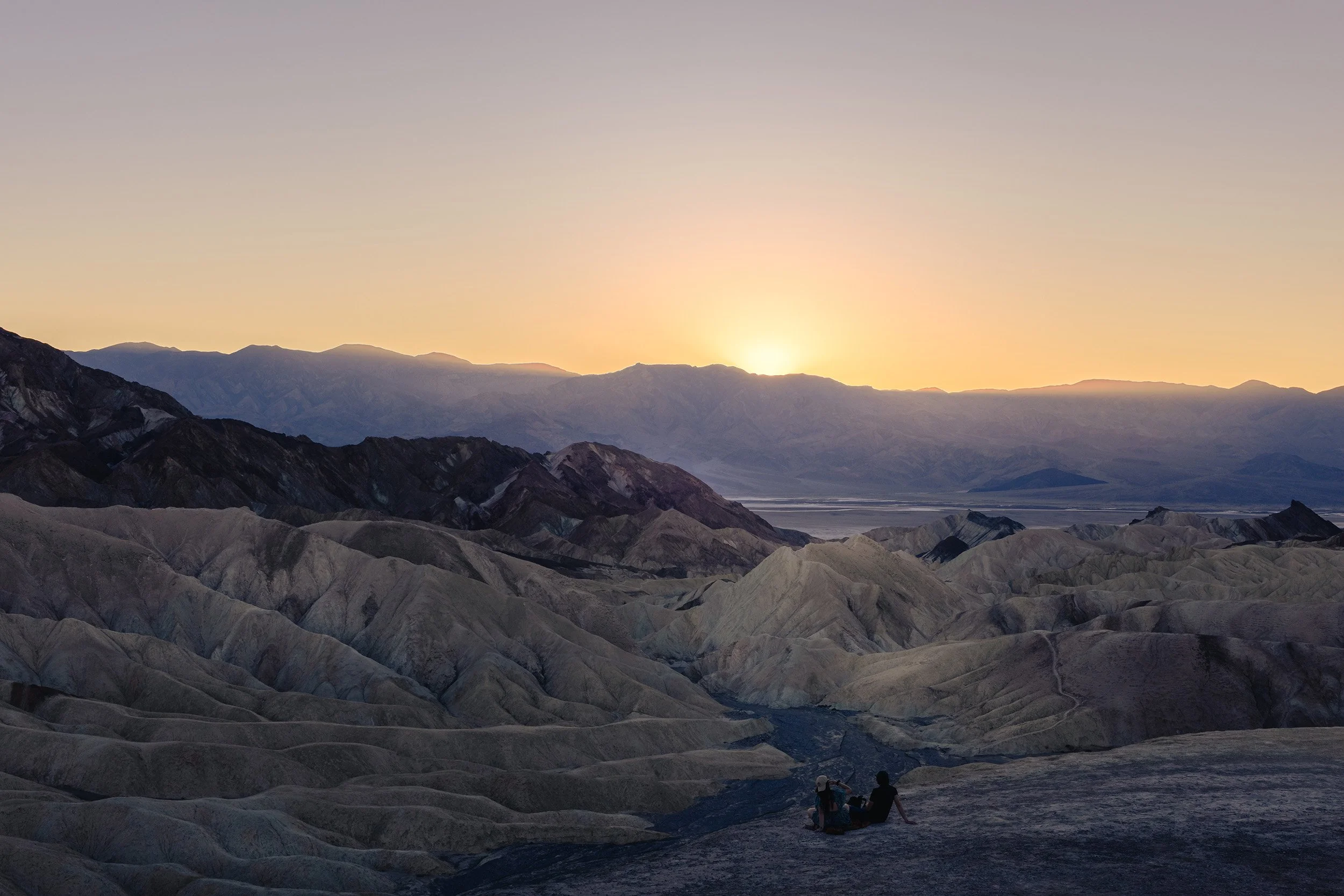 Two people sitting on a rock, watching a sunrise or sunset over a mountainous desert landscape with colorful layered hills and distant mountains.