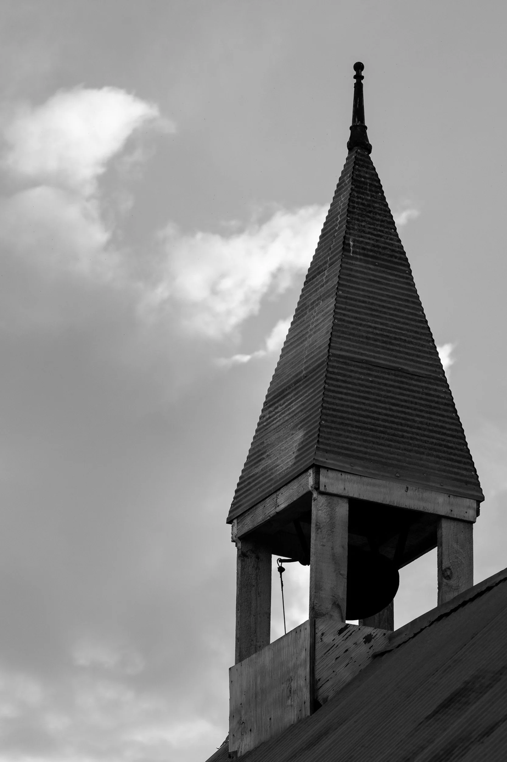 Black and white photo of a church steeple with a weather vane at the top, against a cloudy sky.