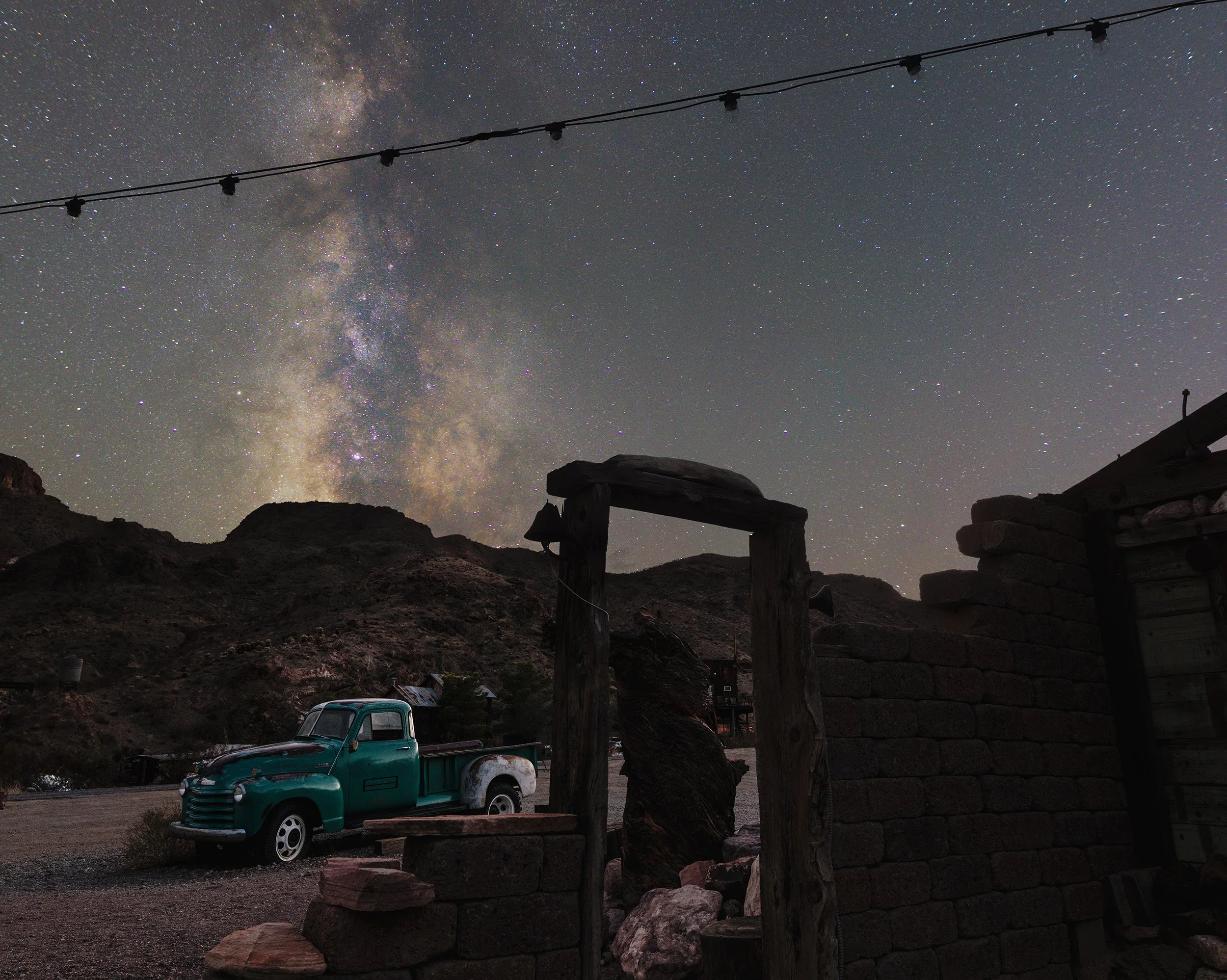 Night sky with the Milky Way galaxy visible, a string of lights overhead, a vintage pickup truck parked near a stone wall, and rugged hills in the background.