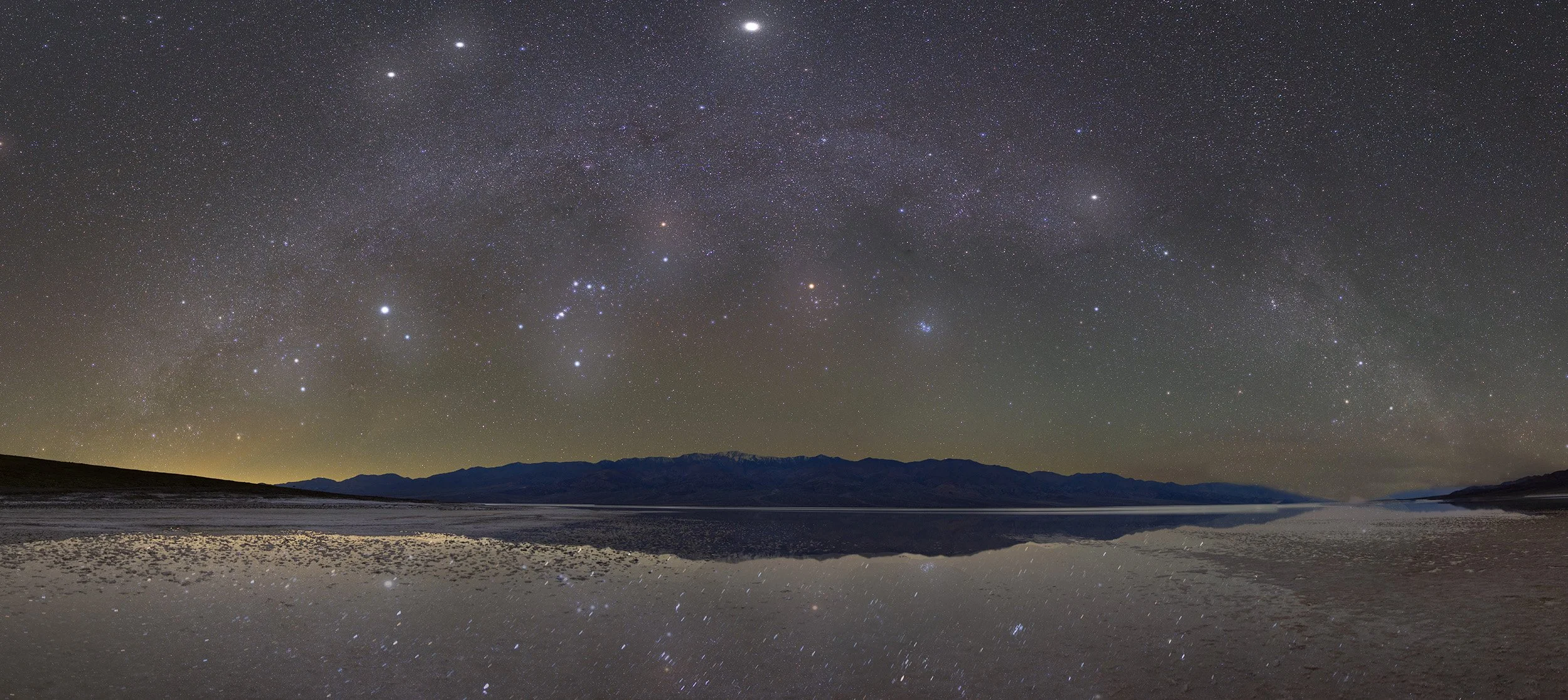 Nighttime landscape with a star-filled sky and the Milky Way, reflected on a calm body of water, with mountains in the distance.