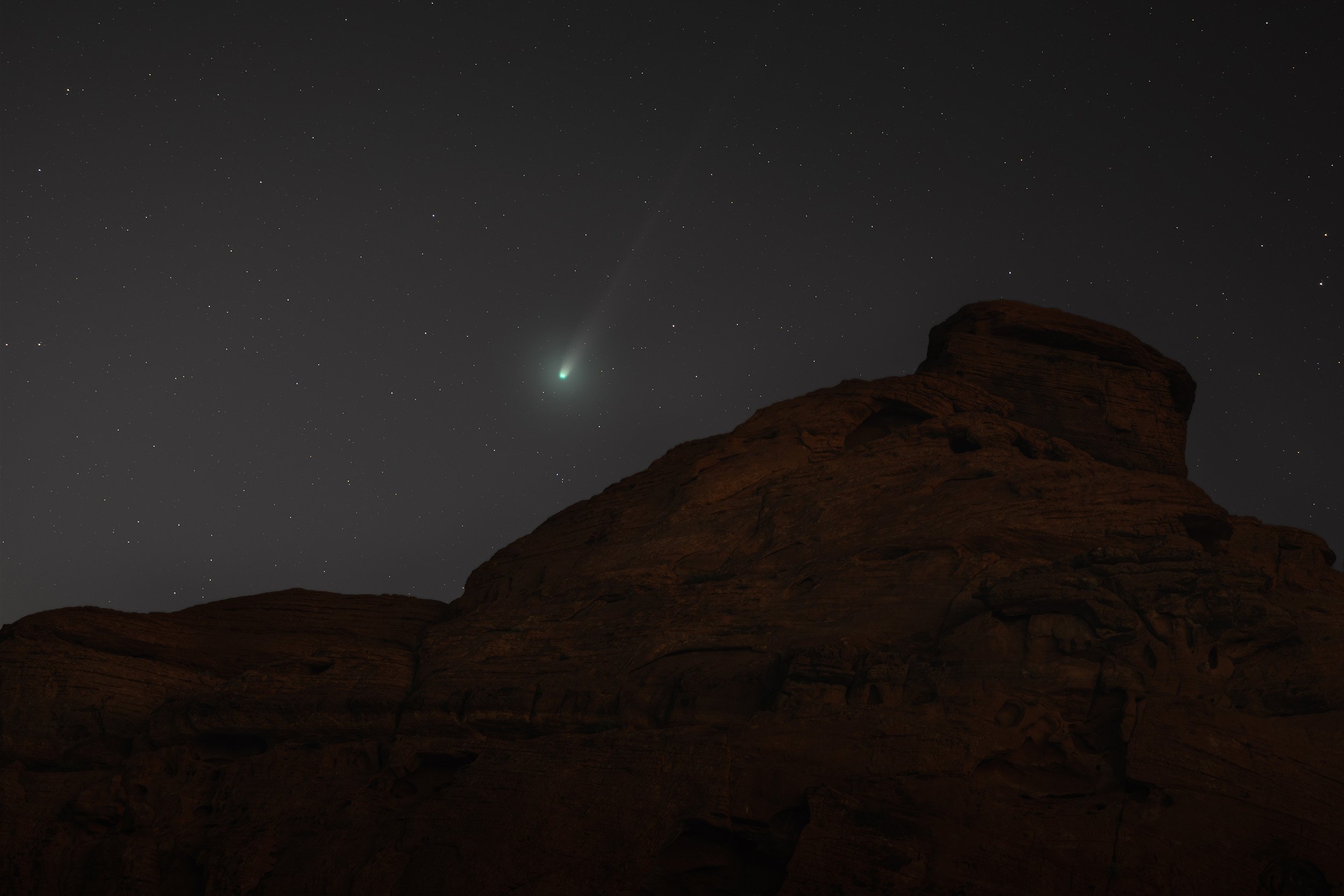 Nighttime desert landscape with a large rock formation silhouette in the foreground, stars scattered across the night sky, and a bright comet or shooting star with a tail visible near the top left.