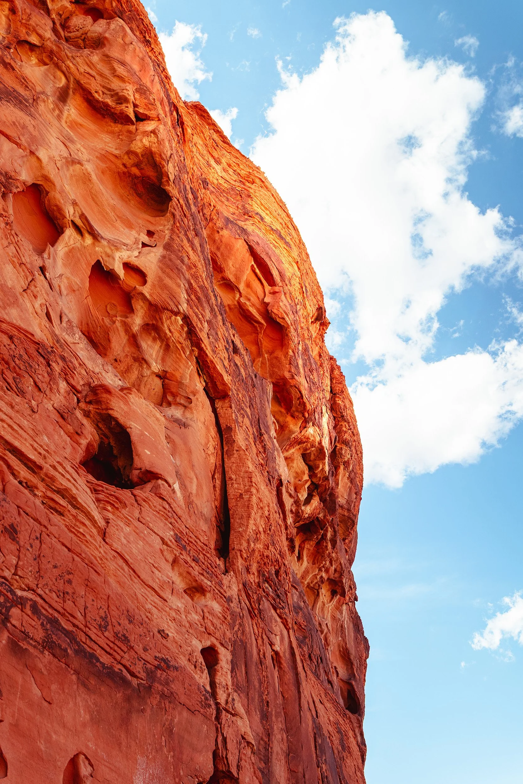 Close-up view of a red-orange sandstone cliff with erosion holes and formations, beneath a partly cloudy blue sky.