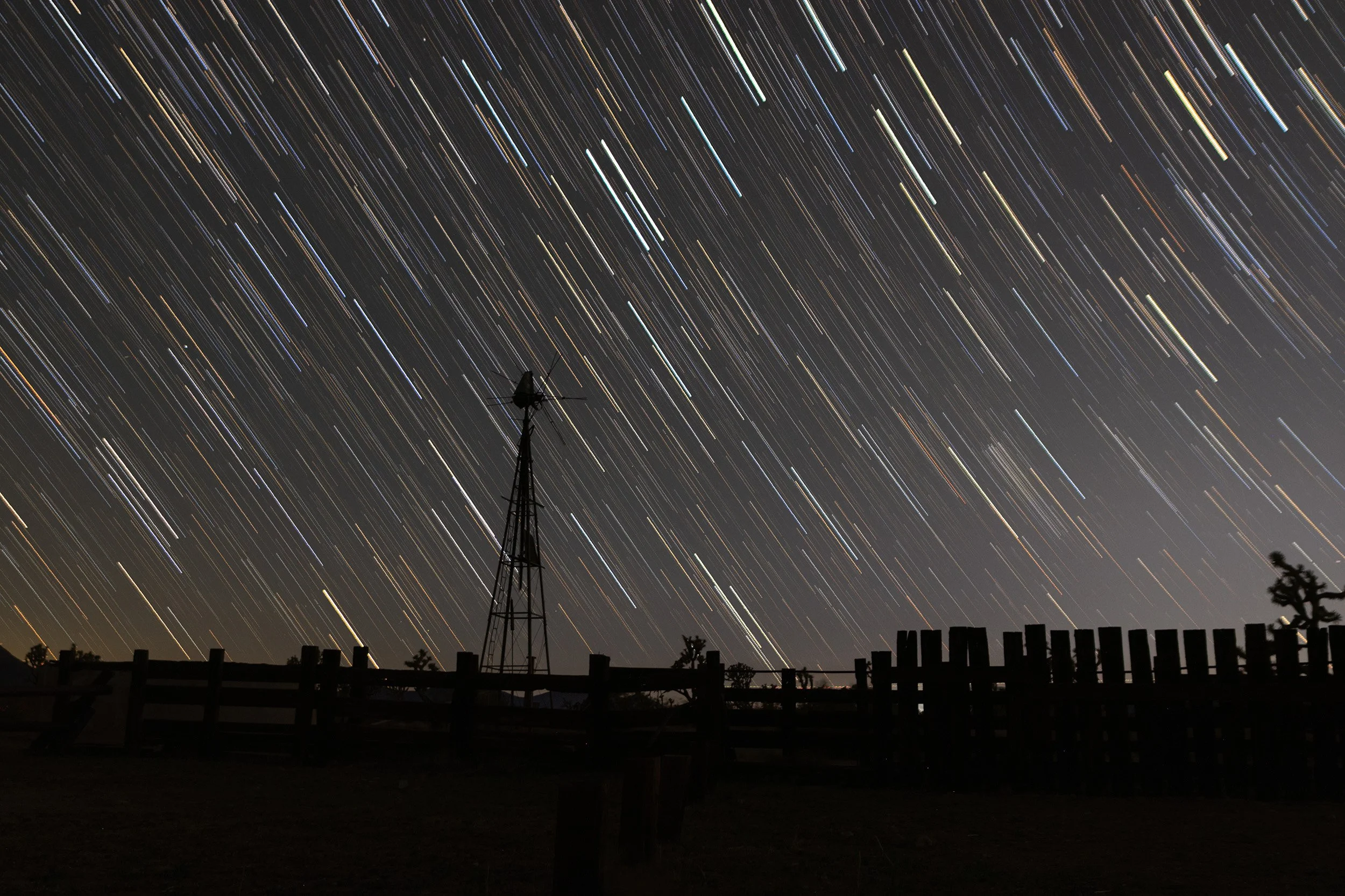 Long exposure photograph of star trails in the night sky over a silhouetted landscape with a weather vane and a wooden fence.