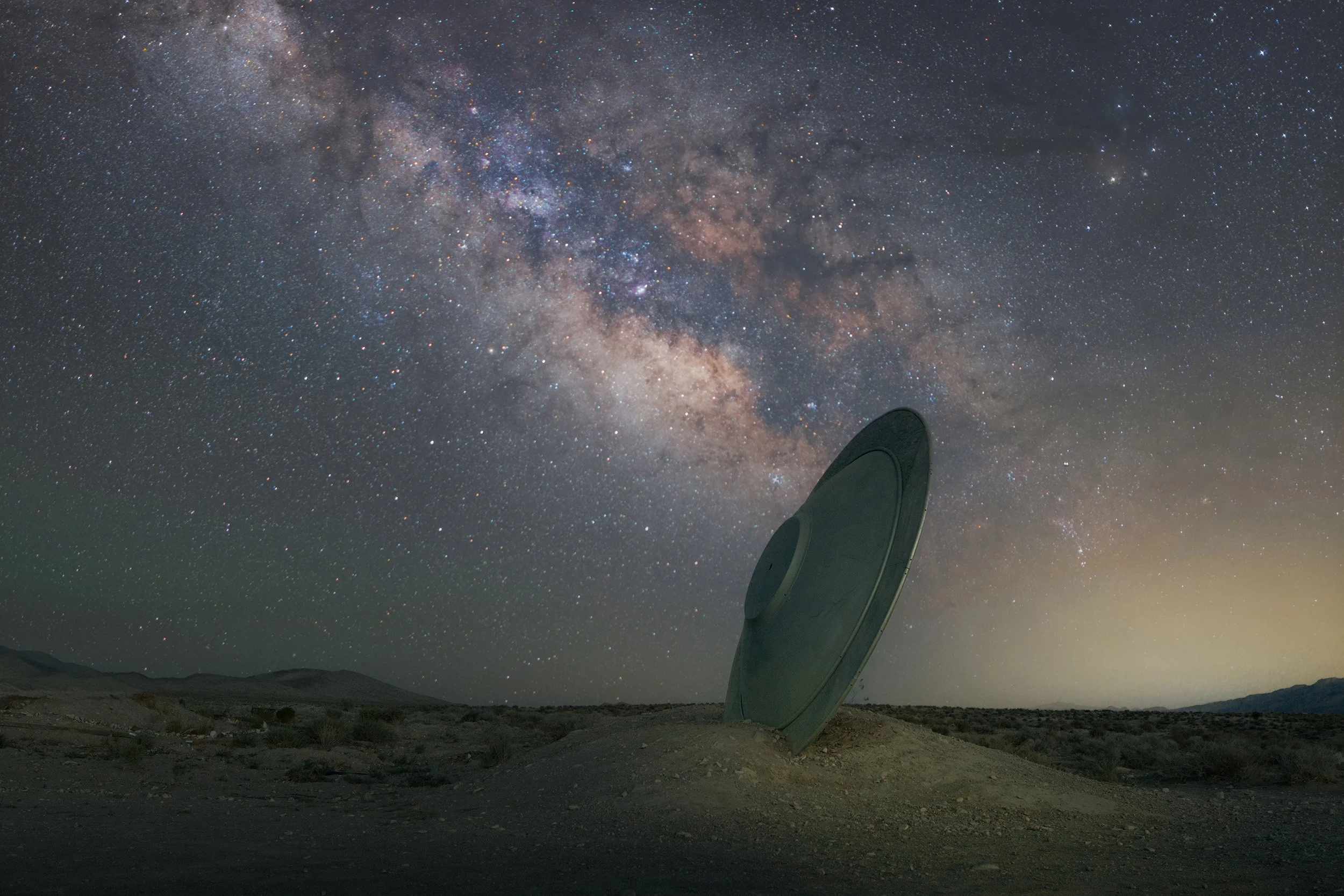 A large radio telescope dish in a desert landscape under a starry night sky with the Milky Way galaxy visible.