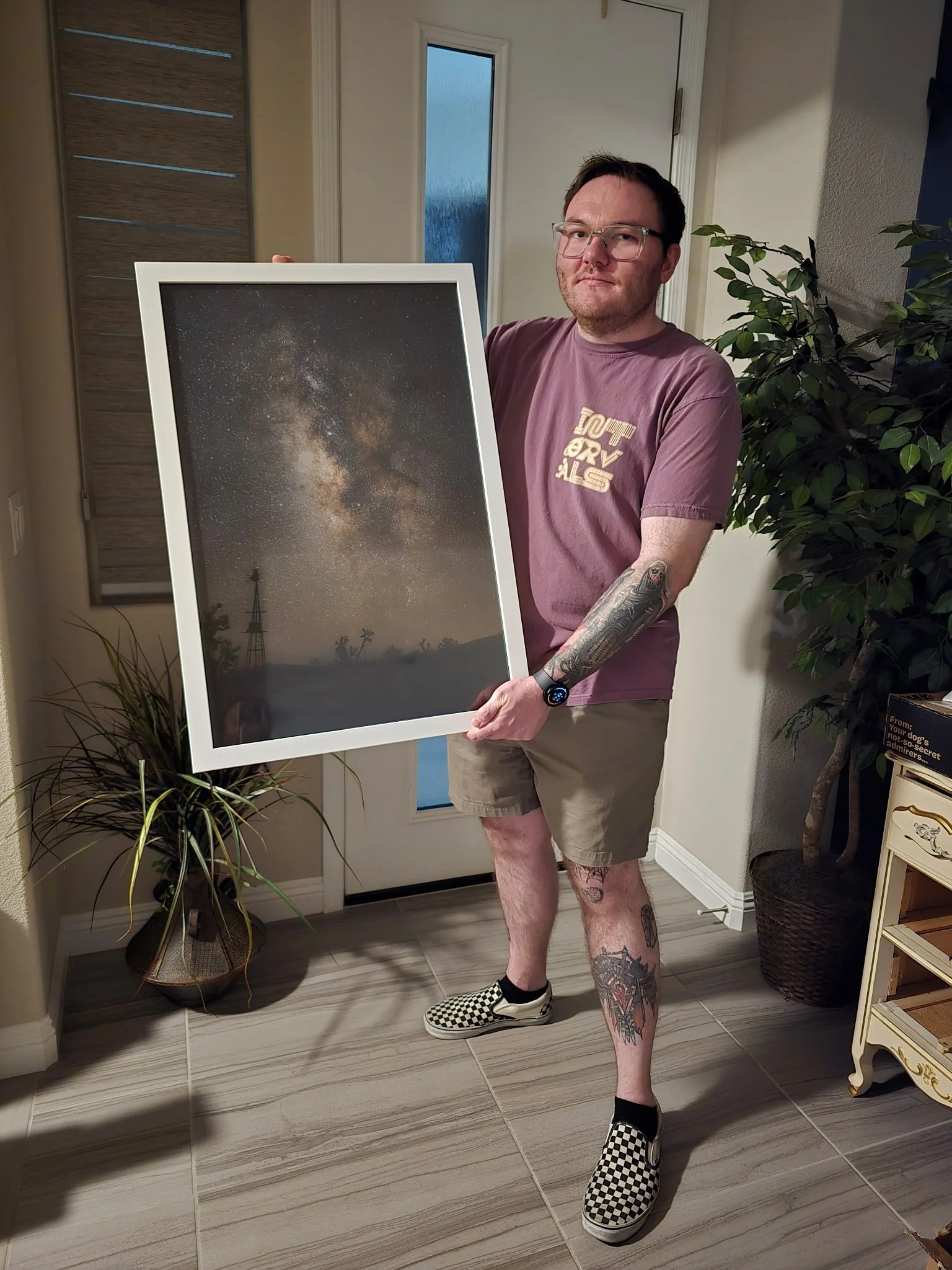 A man with tattoos on his legs and arms, wearing glasses, a maroon T-shirt, beige shorts, and checkered slip-on shoes, is standing indoors holding a framed photo of the night sky with stars and the Milky Way galaxy.