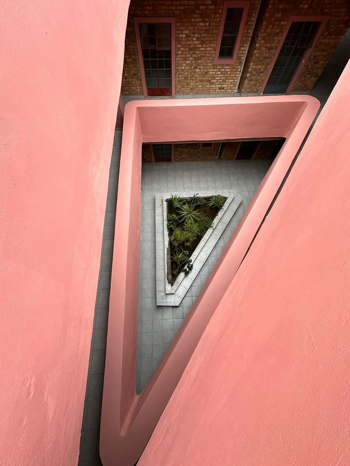 Photo of a pink building courtyard taken from the upper floor, showing a triangular planter with various green plants and surrounding gray tiled ground.
