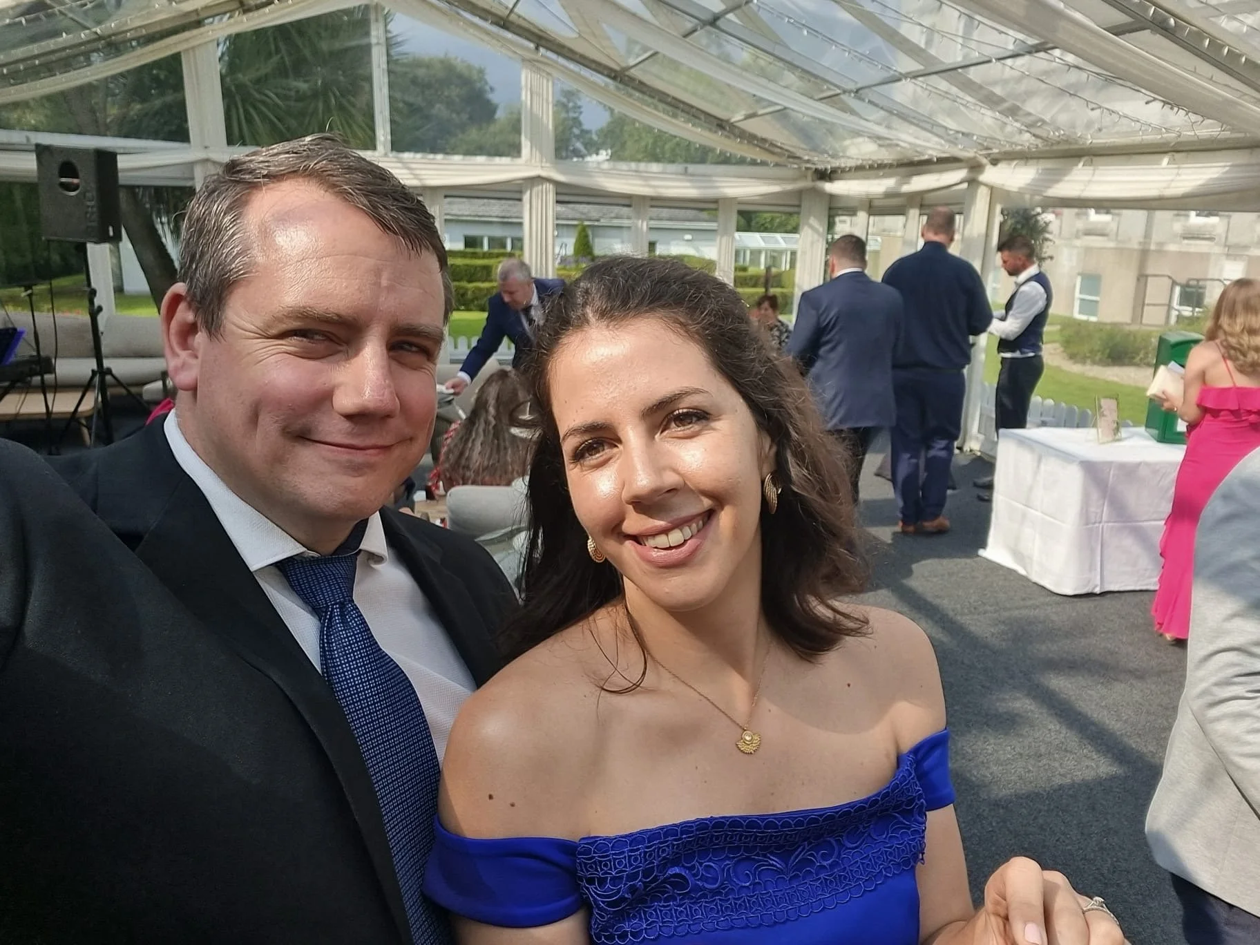 A couple taking a selfie at a formal outdoor event, with guests mingling in the background under a transparent tent on a bright day.