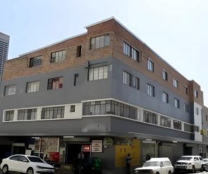 Multi-story building with shops on the ground floor, cars parked in front, and a clear blue sky.