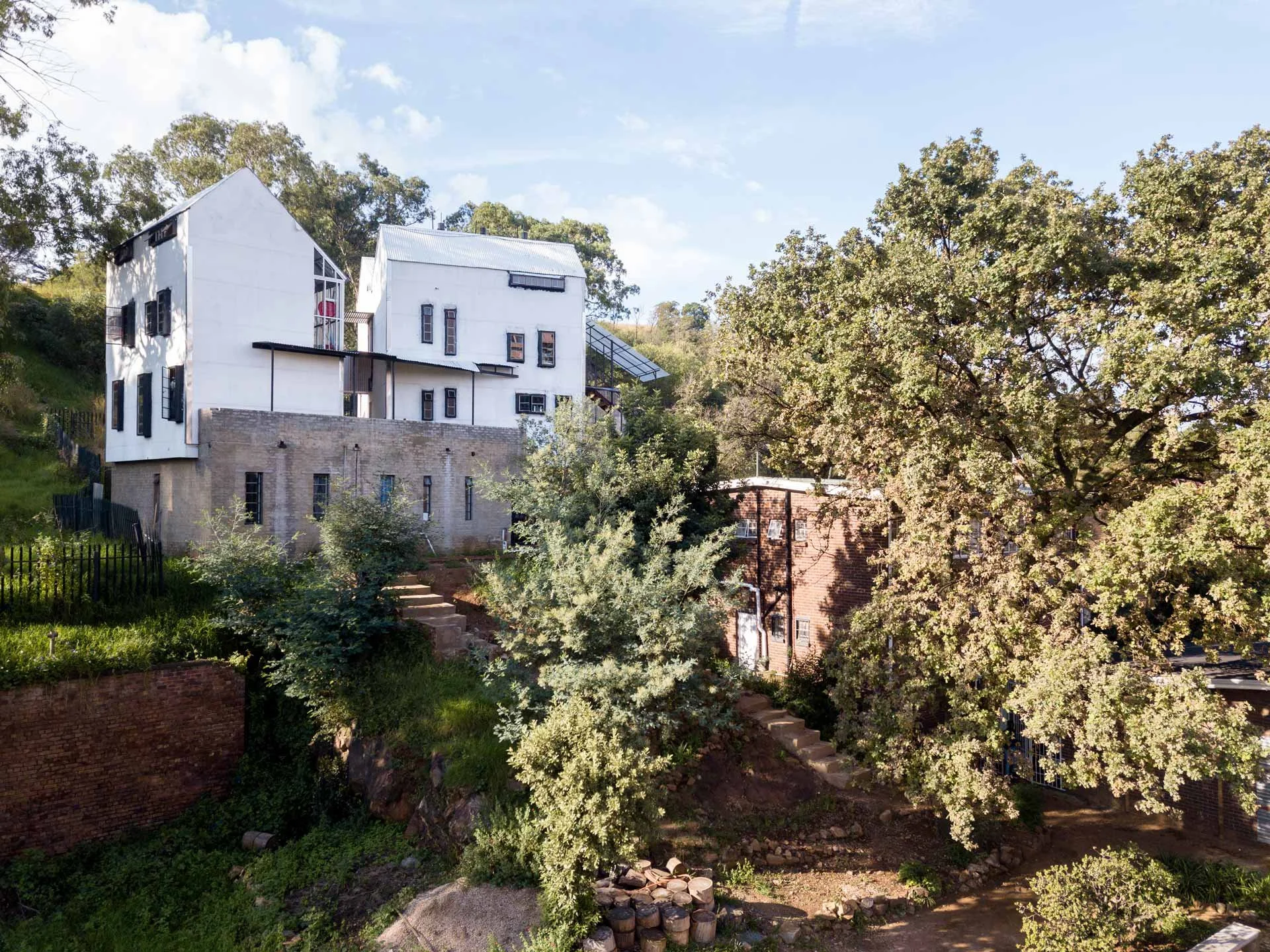 A multi-story house built on a hill with a wooden and concrete exterior, surrounded by trees and greenery, under a partly cloudy sky.