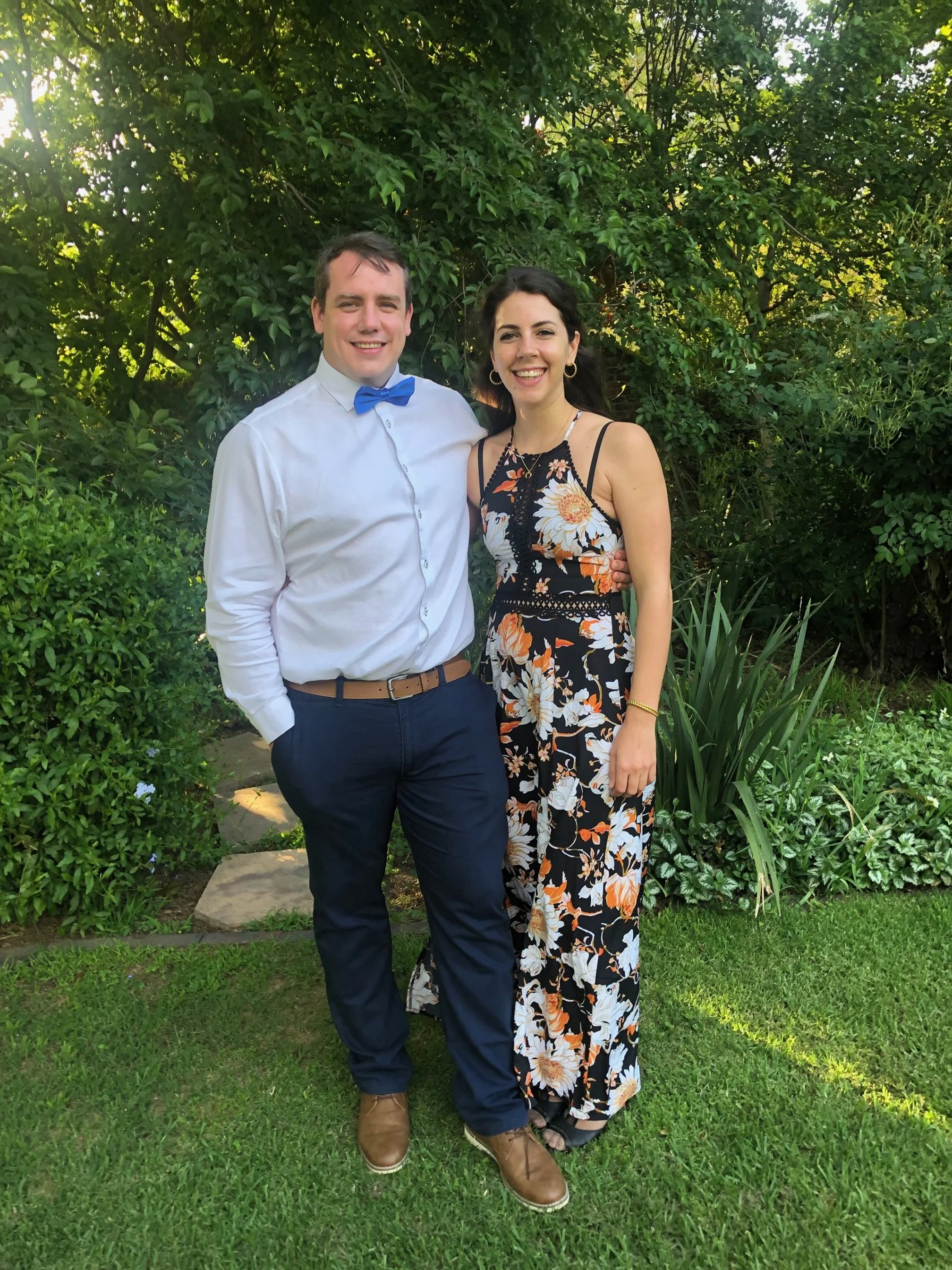 A man in a white shirt with a blue bow tie and navy pants stands with a woman in a black floral dress under a tree in a lush garden.