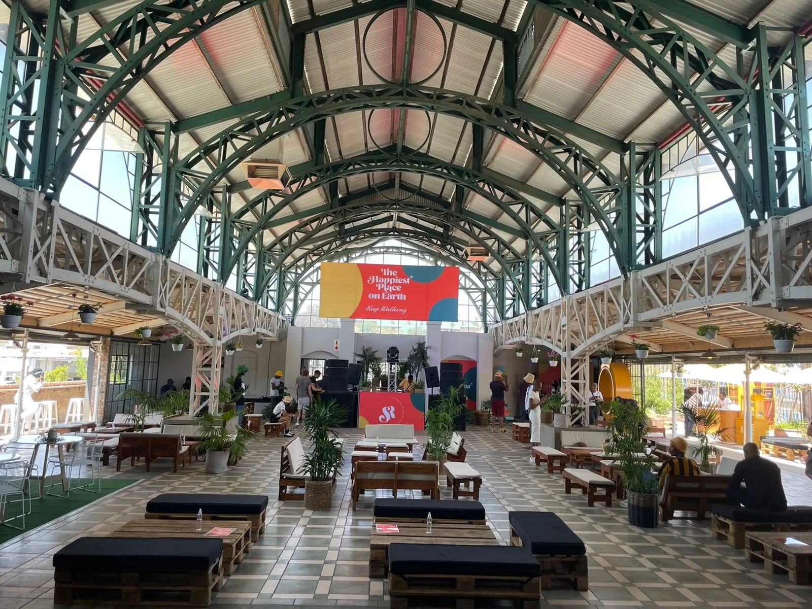 Interior of a spacious, modern event space with a large arched glass ceiling, seating areas with wooden benches and plants, a stage with a large screen displaying 'The Happiest Place on Earth,' and people setting up or preparing for an event.