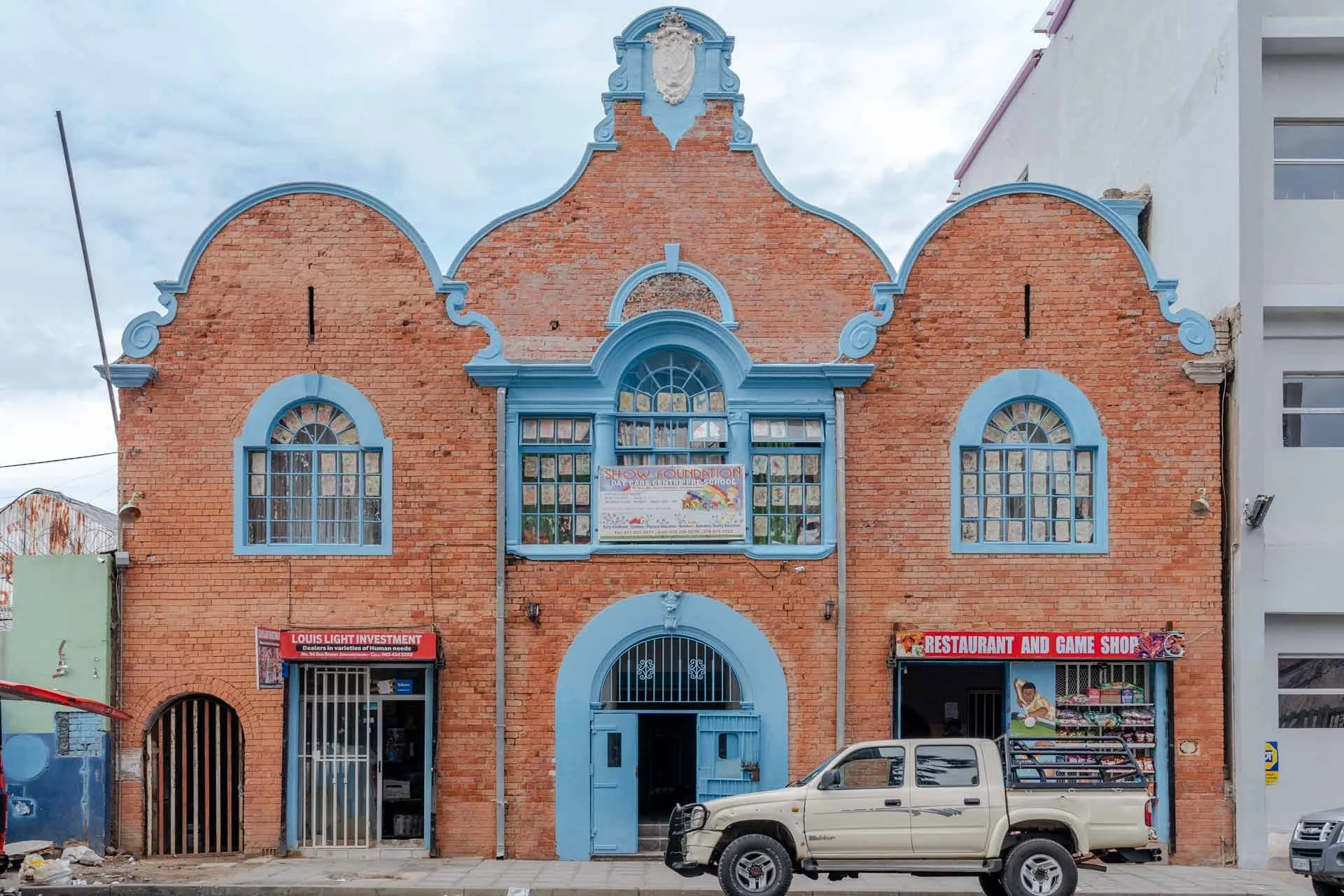 A two-story brick building with blue architectural details and arched windows, with signs indicating it is a restaurant, game shop, and investment business. There is a beige pickup truck parked in front of the building.