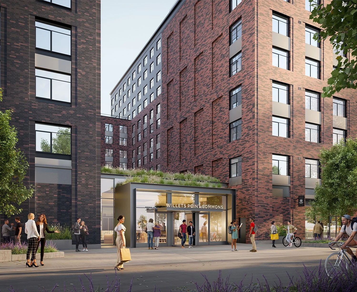 People walking and riding bikes outside a modern, multi-story brick apartment building with an entrance sign reading 'Willets Point Commons'.