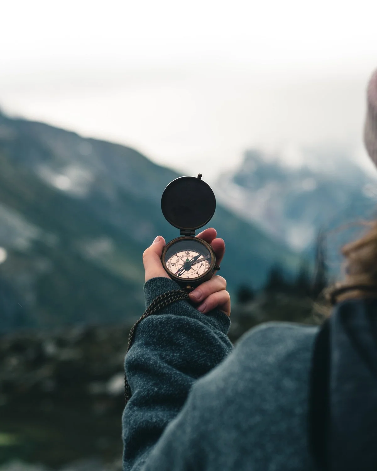 Person holding a compass in a mountainous outdoor setting with snow-capped peaks in the background.