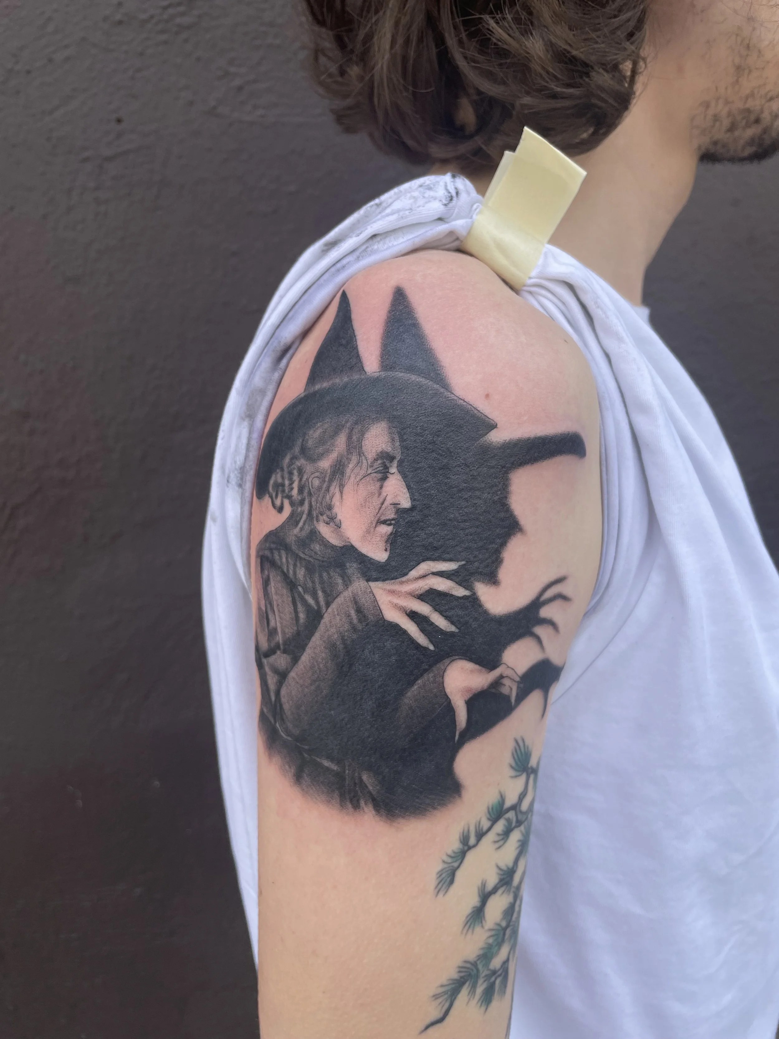 Tattoo of a witch with a black pointed hat, with a background of a full moon and silhouettes of flying bats or crows.