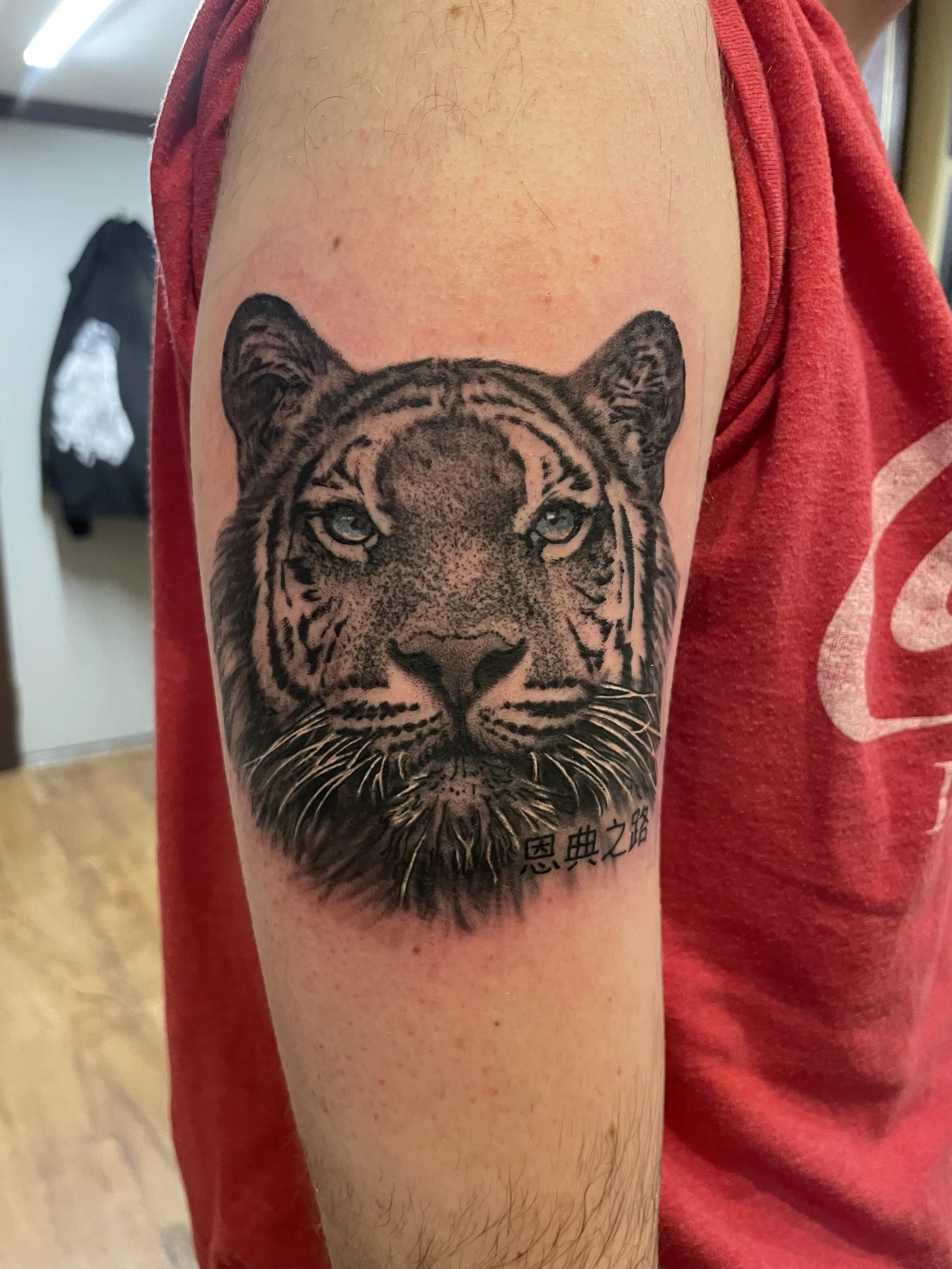 Tattoo of a tiger's face with piercing eyes on a person's upper arm, with Chinese characters below the tiger's chin.