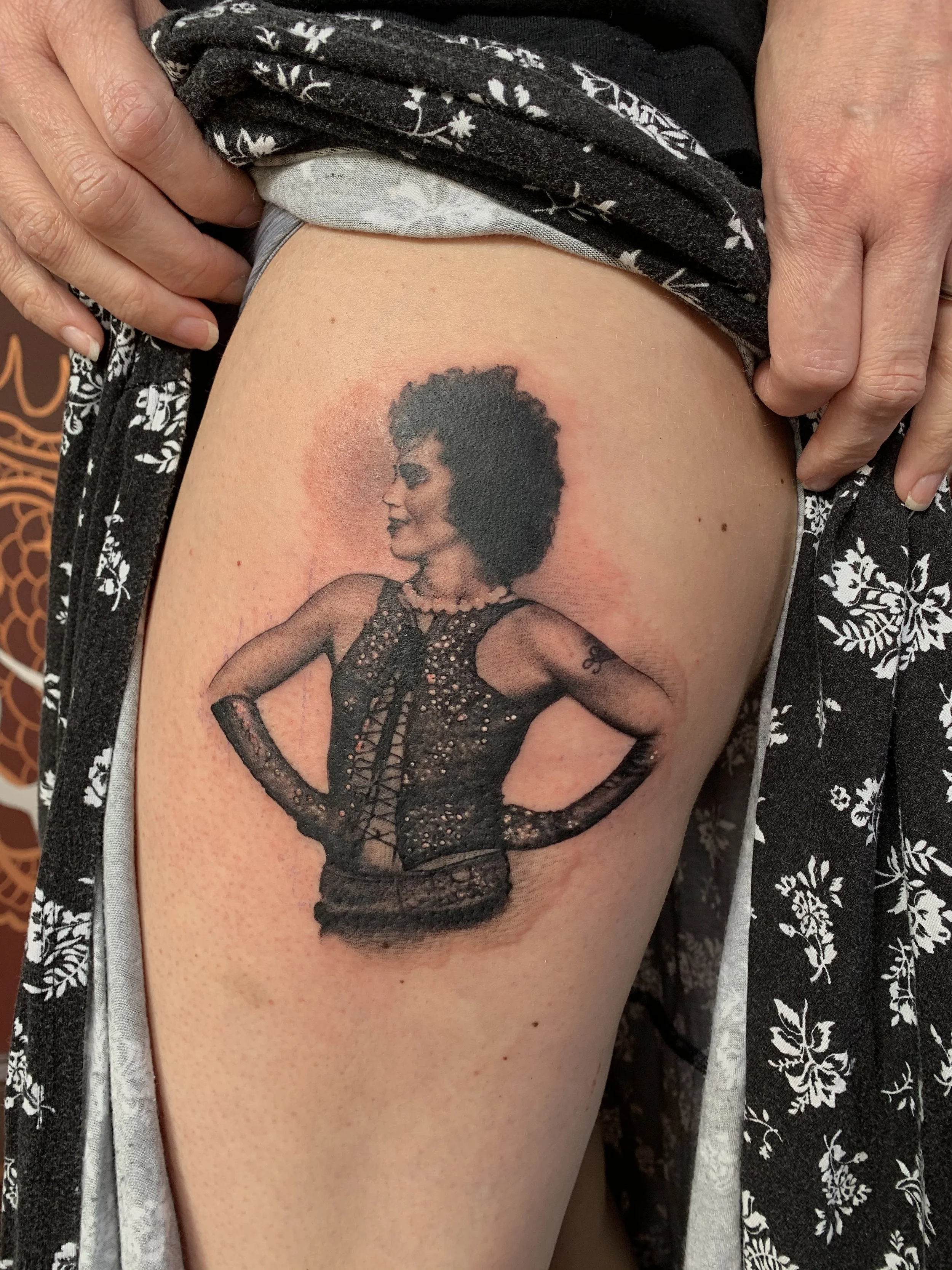 Tattoo of a woman in vintage clothing with her hands on her hips, wearing sunglasses and a lace top, on a person's upper arm.