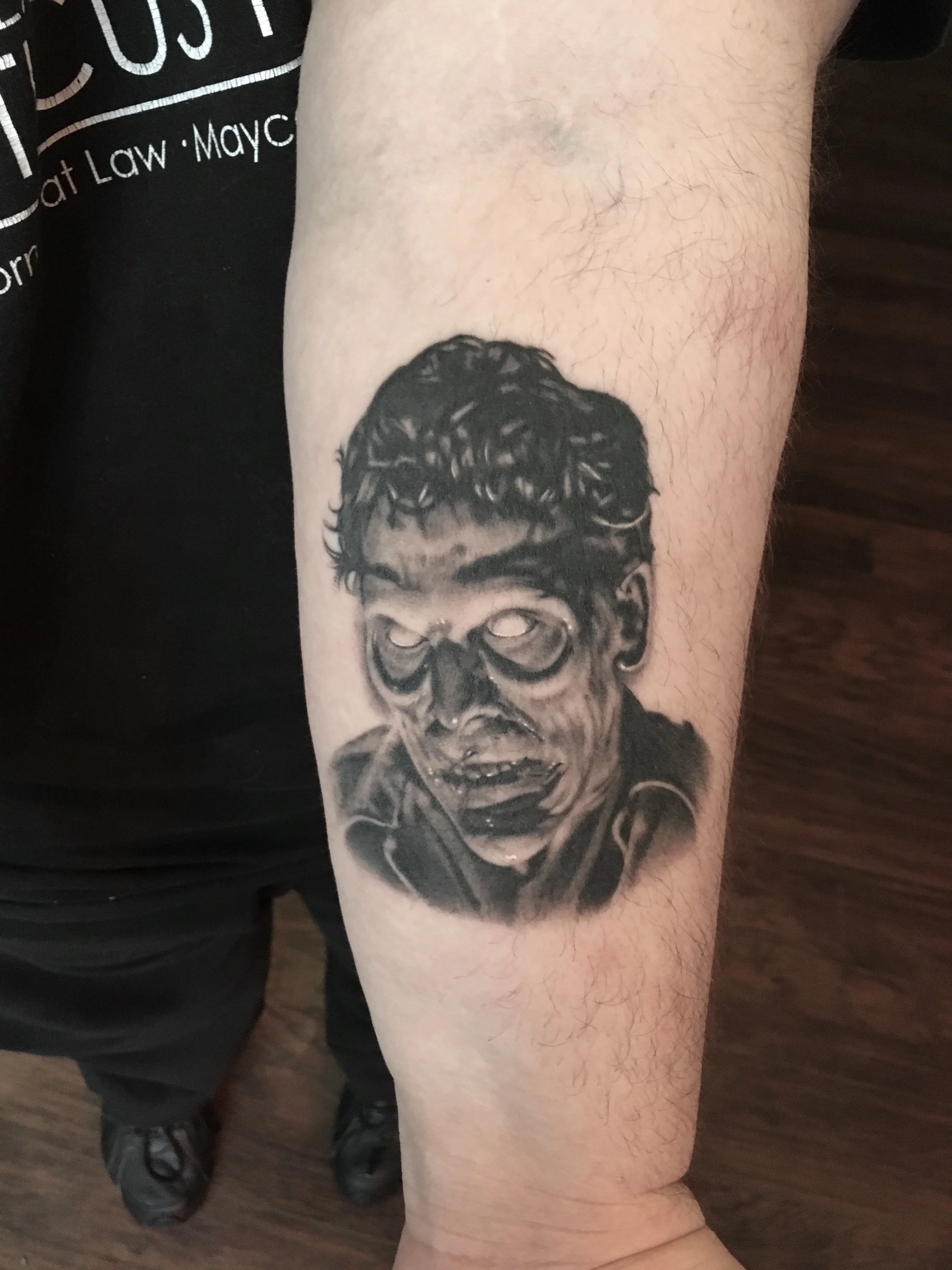 Black and gray tattoo of a zombie or undead figure with hollow eyes, disheveled hair, and a damaged face, on a person's forearm.