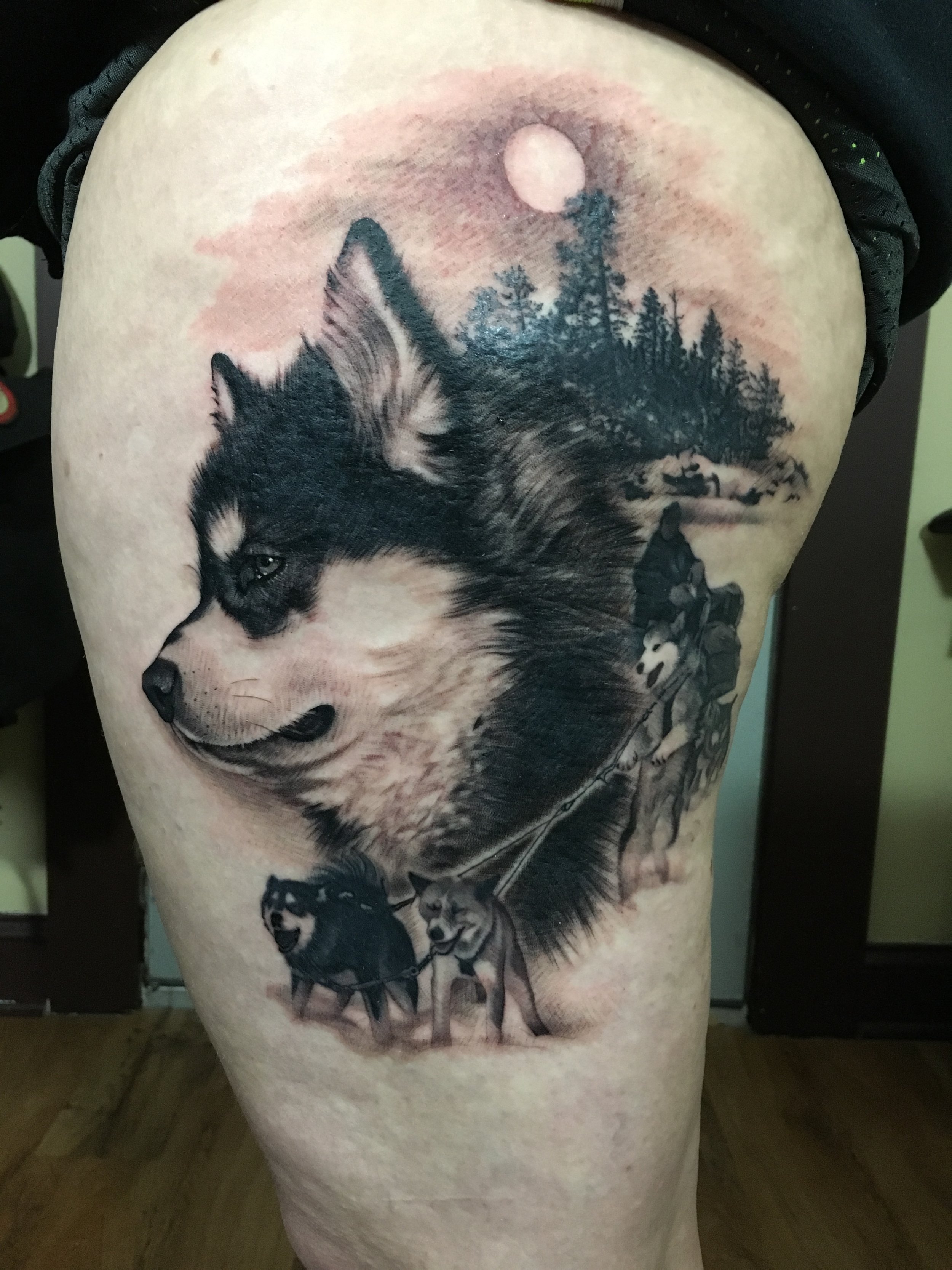 Tattoo of a husky dog with a forest background, moon, and wolves on the person's thigh.