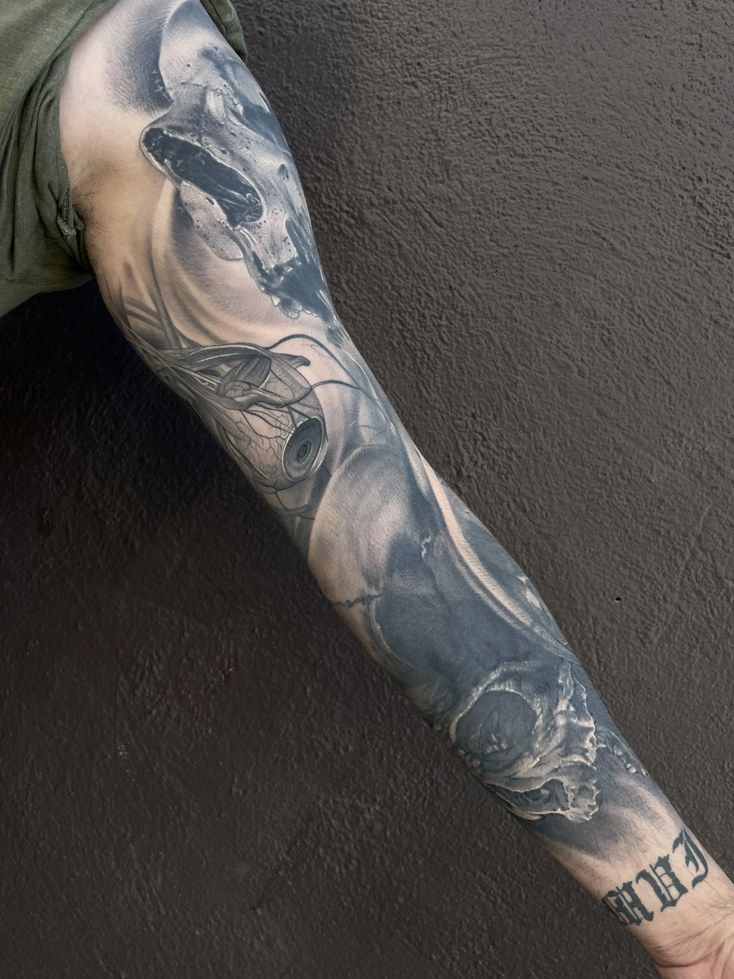 Close-up of a tattoo sleeve on a person's arm, featuring a skull, an eye with detailed shading, and various abstract and realistic elements in black and gray ink.