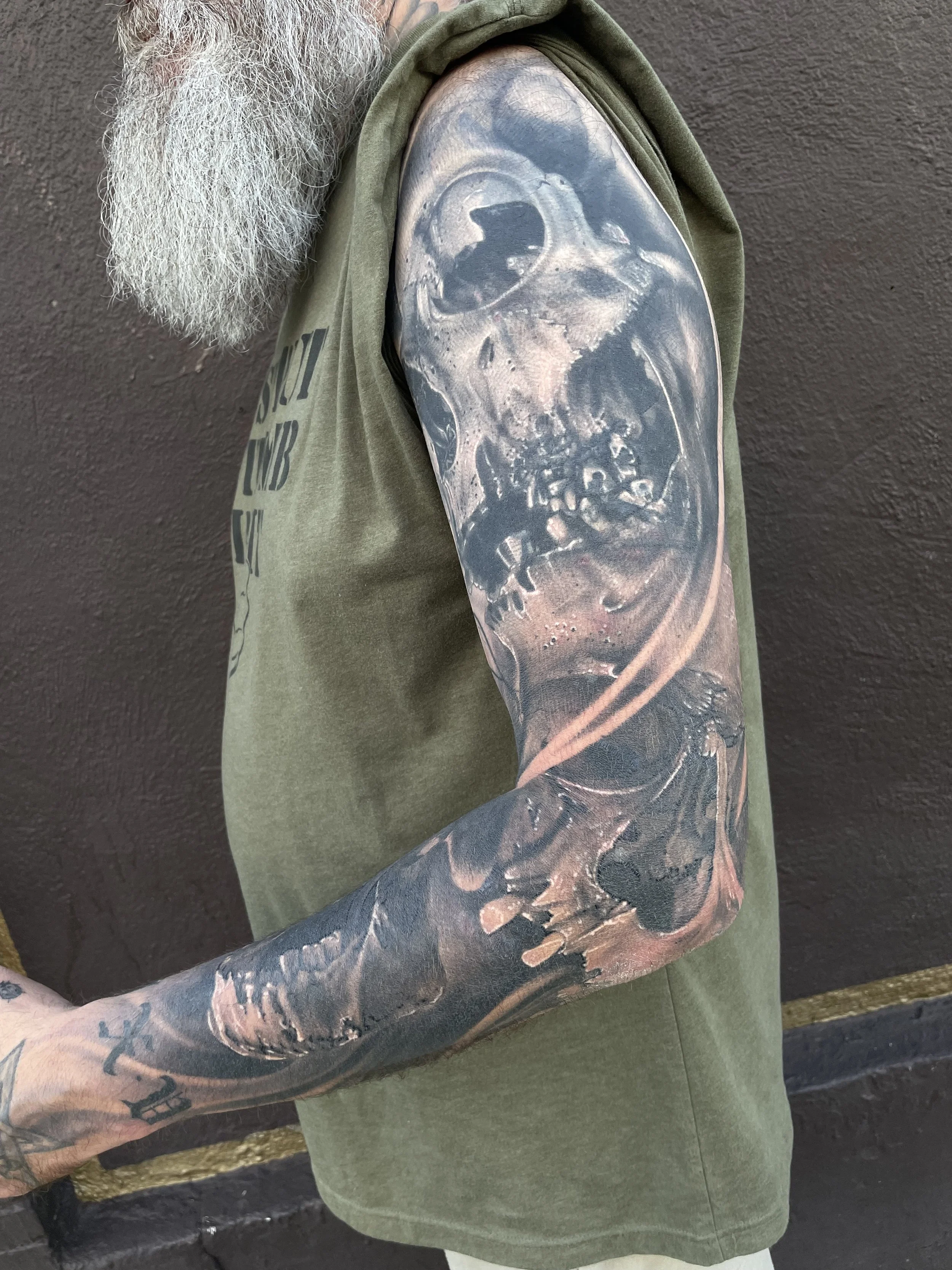 A person with a long white beard wearing a green sleeveless hoodie, displaying a detailed black and gray skull tattoo on their upper arm and forearm.