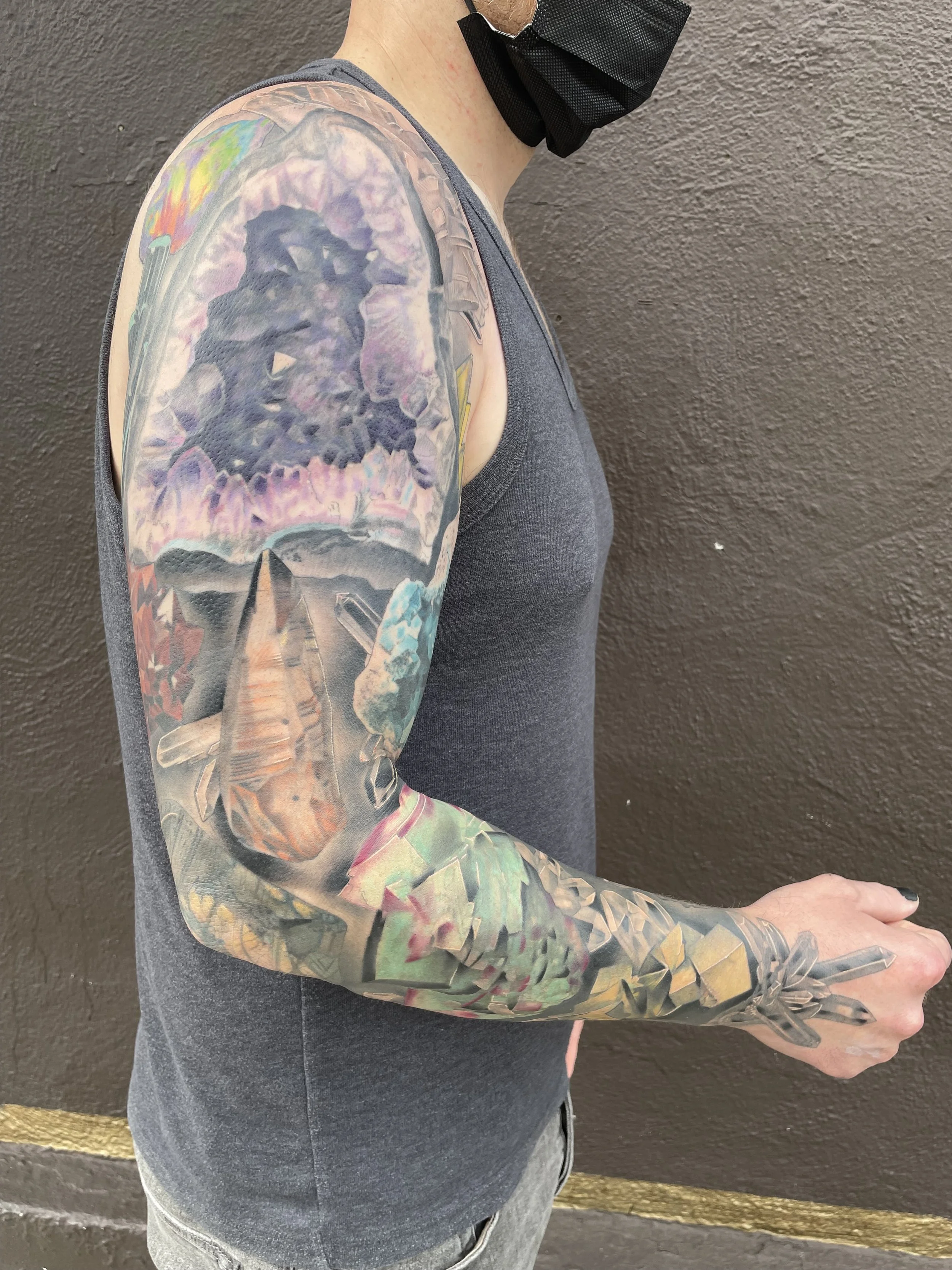 Colorful full sleeve tattoo on a person's arm, featuring a mountain landscape with a cloudy sky and various abstract elements, alongside a black face mask and gray tank top.
